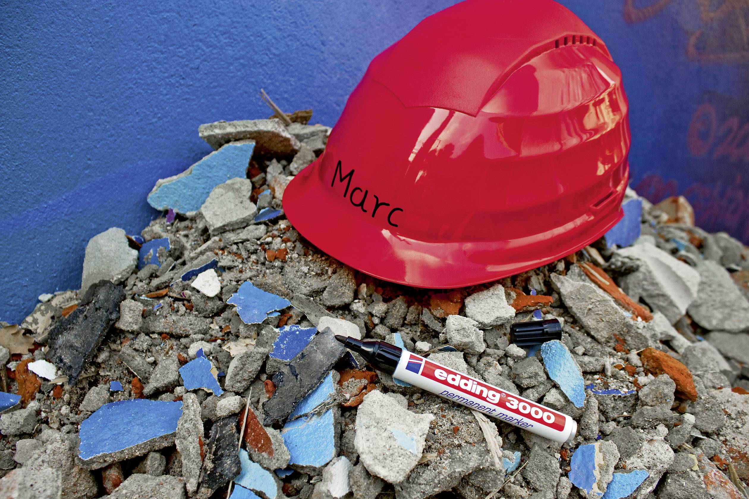 Roter Bauhelm mit Name Marc, liegt auf einem Trümmerhaufen. Daneben ein Marker, Marke edding 3000. Hintergrund: blaue Wand.
