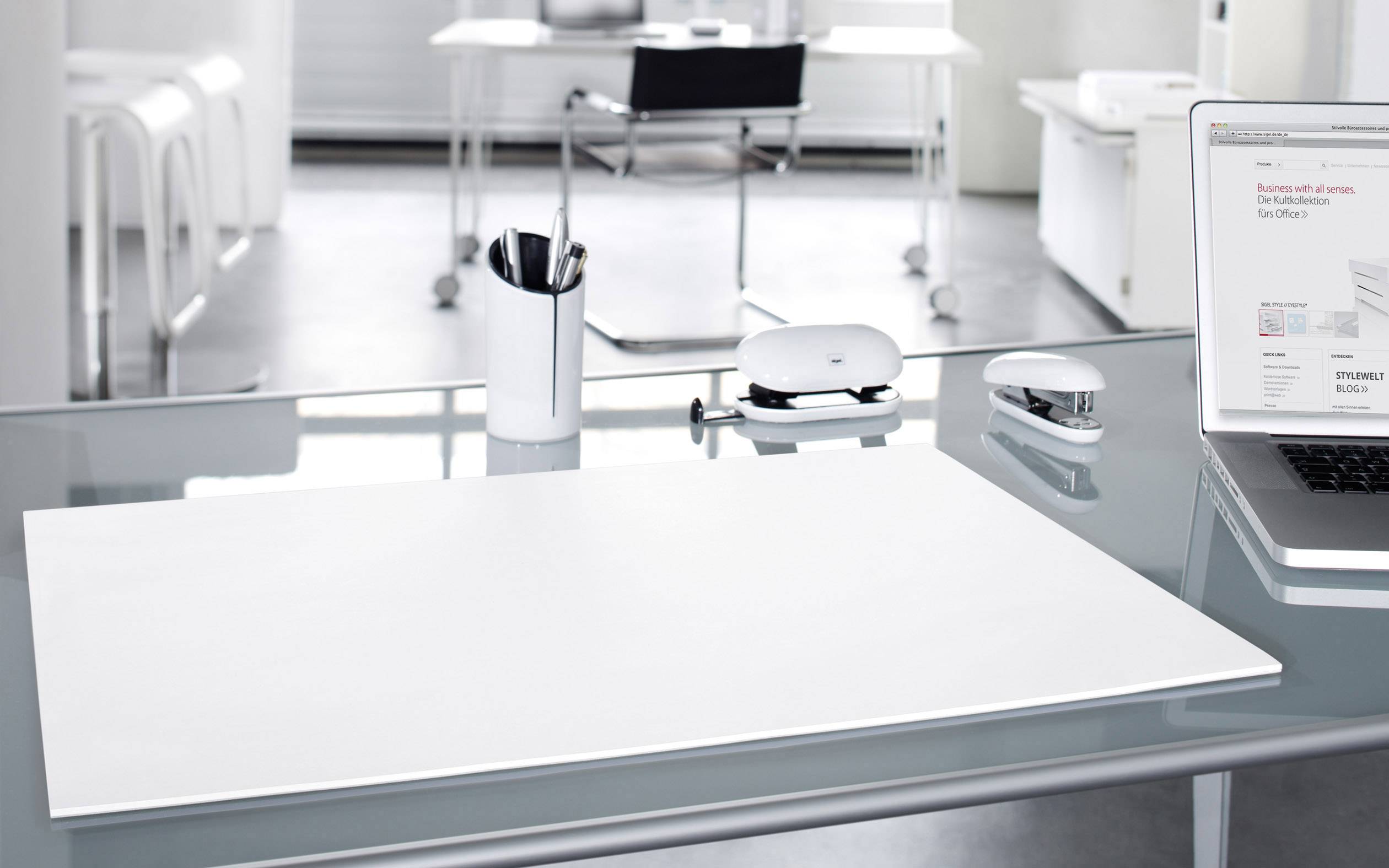 Ein moderner, aufgeräumter Büroraum mit einem Glasbüro-Schreibtisch, auf dem Bürobedarf steht. Im Hintergrund steht ein leerer Stuhl.