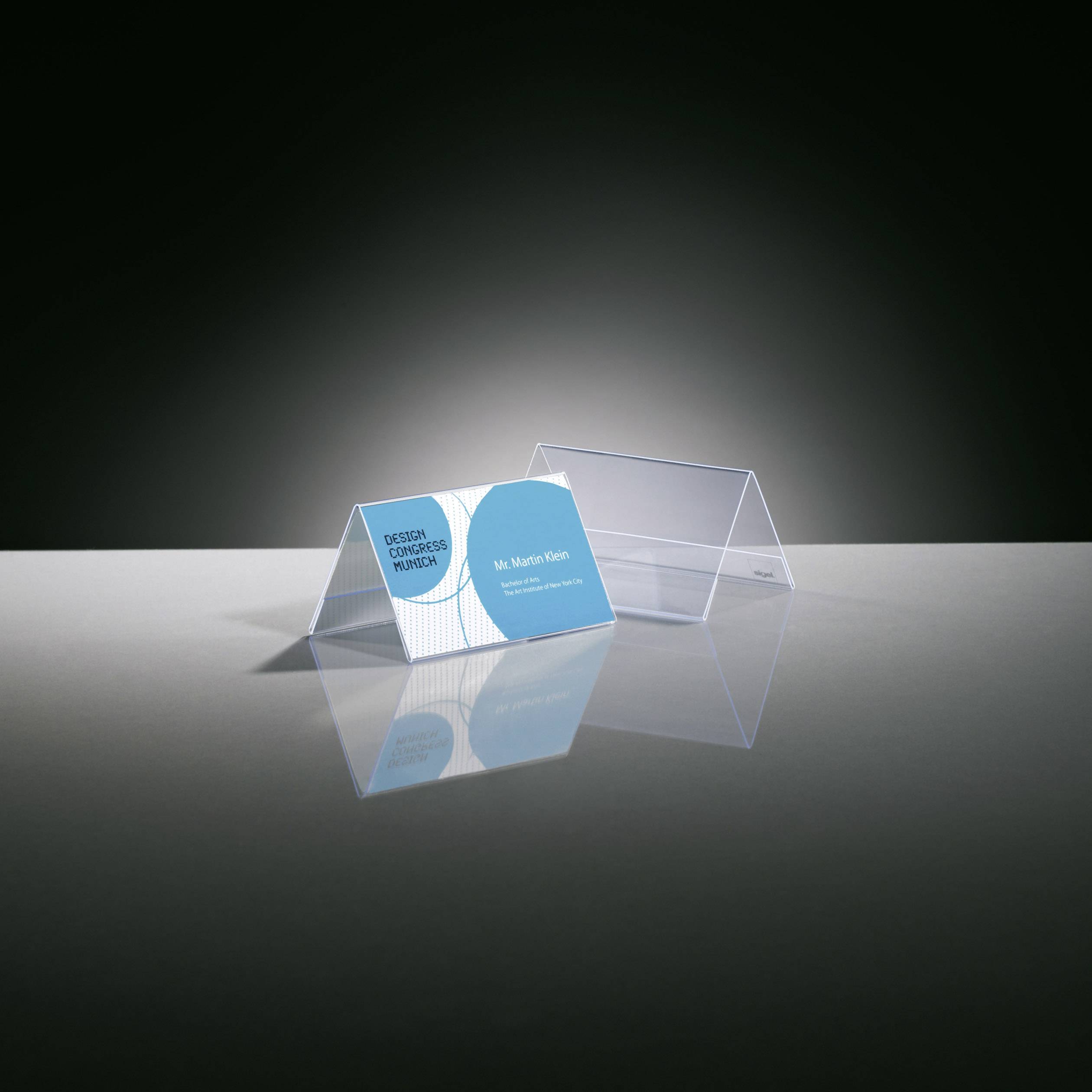 Zwei durchsichtige Visitenkartenhalter auf einem Tisch, der linke enthält eine Karte mit der Aufschrift 'Device for boarding, Designed by Helmut Esslinger' und dem Namen 'Mr. Martin Klein'.