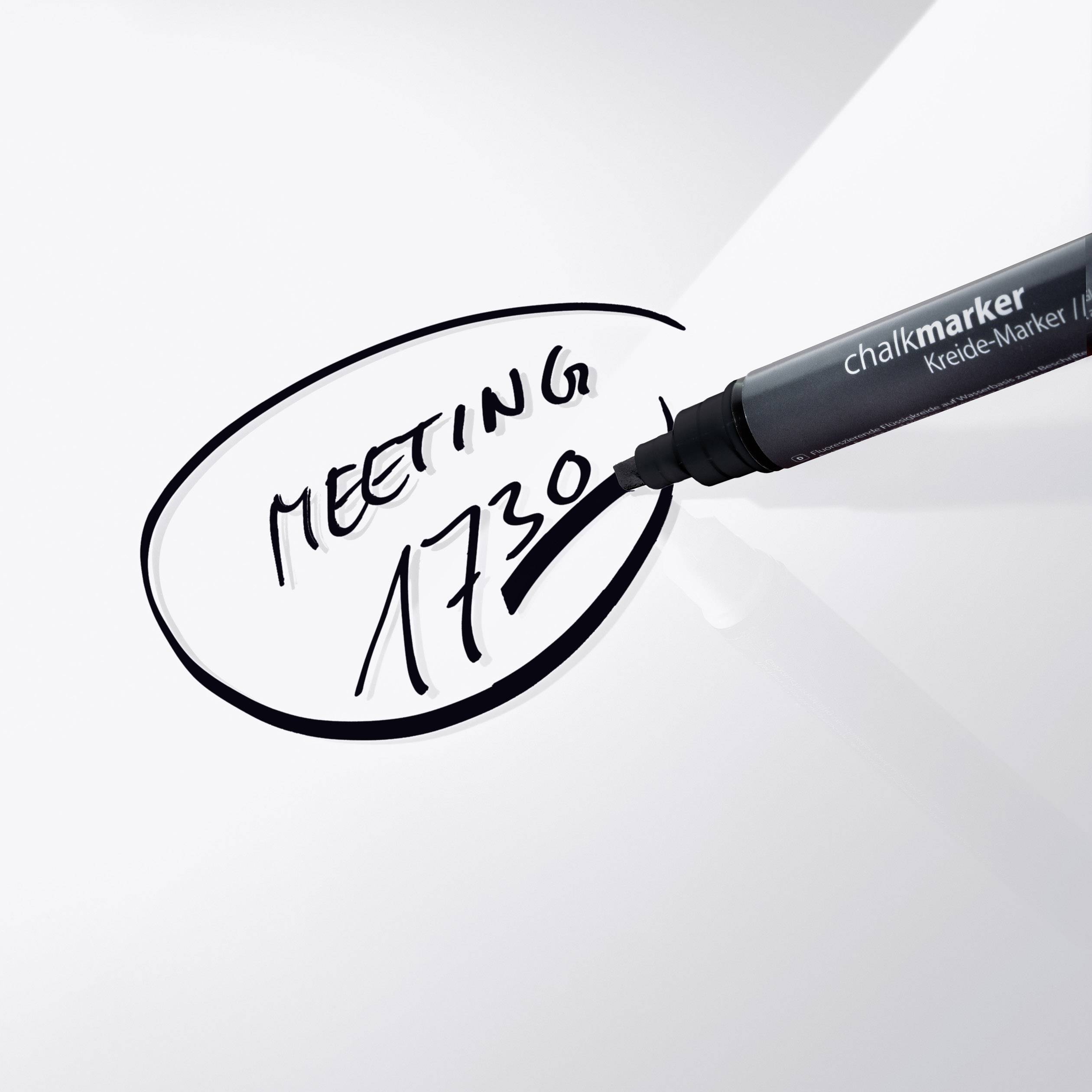 Ein schwarzer Kreidemarker schreibt 'MEETING 17:30' auf eine weiße Oberfläche.