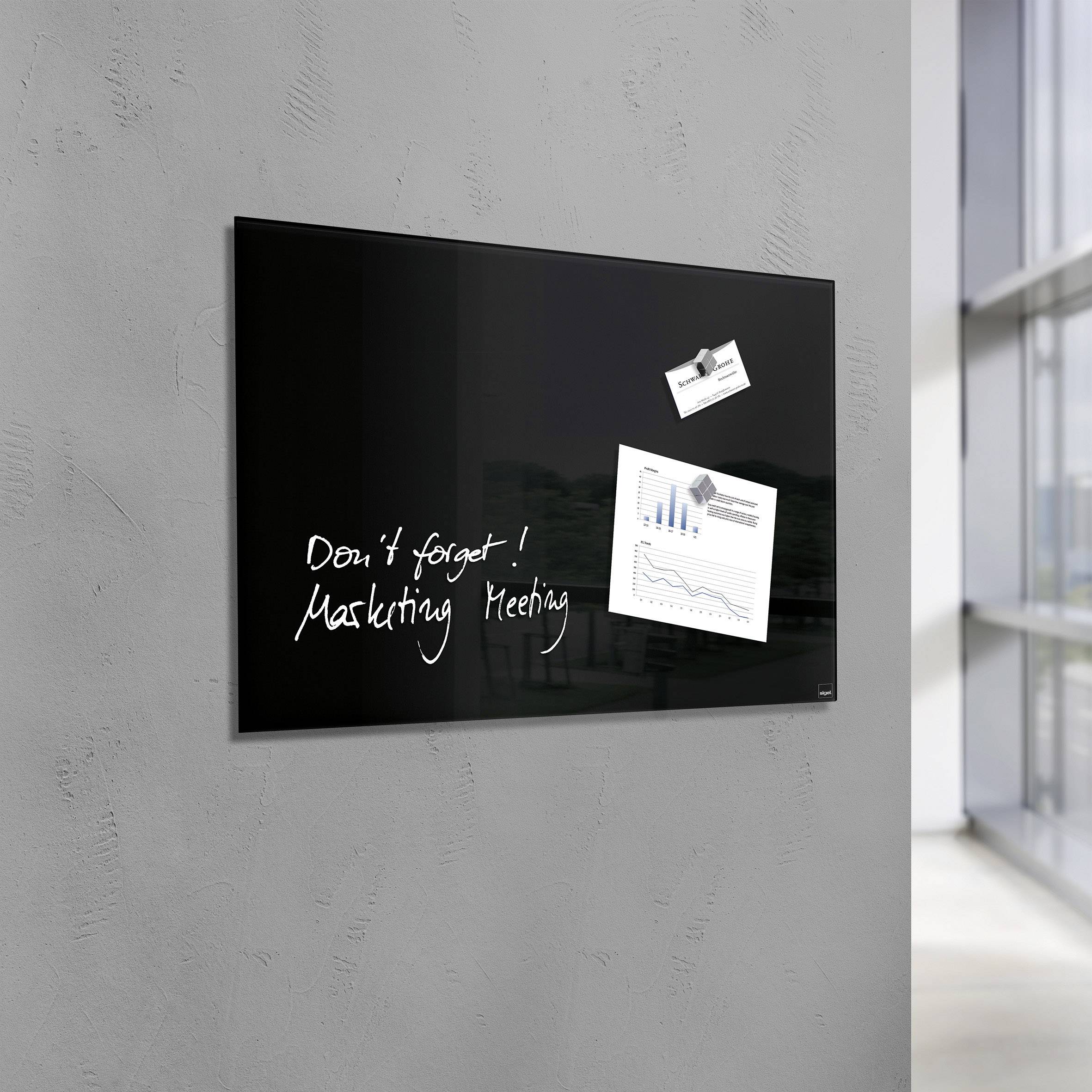 Schwarzes Whiteboard an einer grauen Wand mit der handschriftlichen Notiz 'Don't forget! Marketing Meeting', daneben zwei Grafiken befestigt.