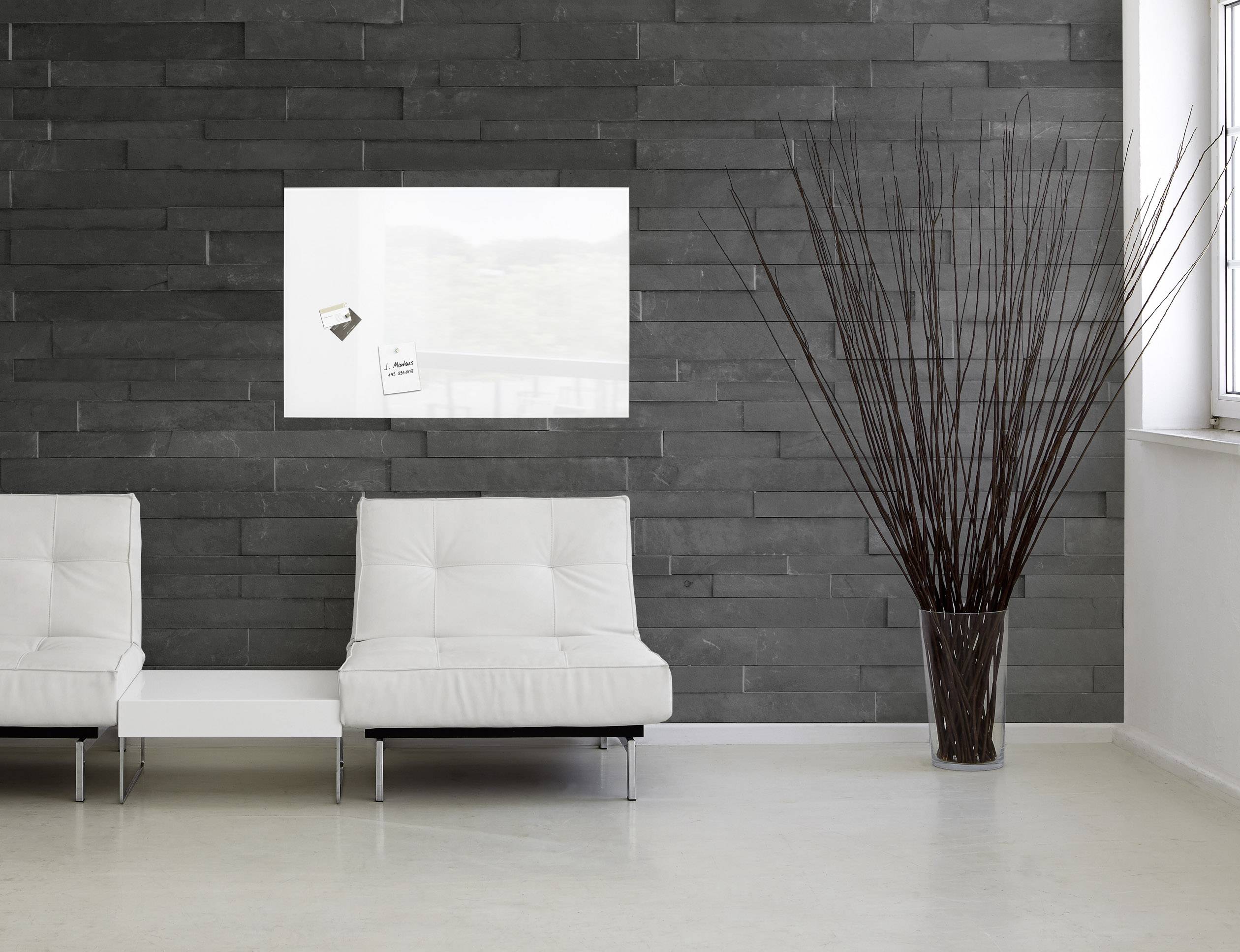 Heller Raum mit zwei weißen Sesseln vor einer dunklen Steinwand. Ein rechteckiges Kunstwerk und ein großer Bodenvase mit Zweigen sind zu sehen.