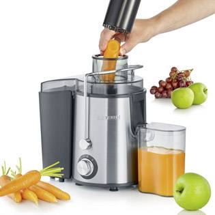 Eine Hand presst eine Karotte in einen Entsafter. Ein Krug fängt den Saft auf. Daneben liegen Karotten, grüne Äpfel und Trauben.