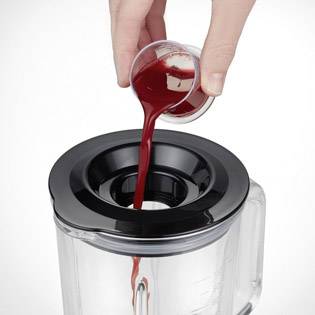 Hand gießt rote Flüssigkeit aus einer kleinen Kanne in einen offenen Mixer.