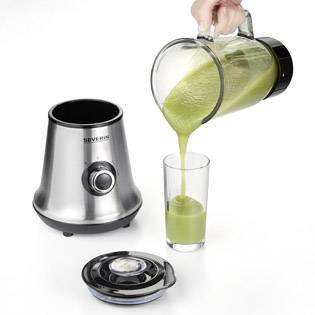 Eine Hand gießt grünen Smoothie aus einem Mixerkrug in ein Glas. Der Mixer steht auf dem Tisch mit einem abgenommenen Deckel.