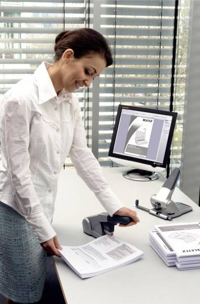 Eine Frau im Büro scannt Dokumente an einem Scanner. Ein Computerbildschirm zeigt ein geöffnetes Dokument. Im Hintergrund sind Fensterjalousien.