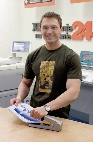 Eine Person steht lächelnd an einem Drucker in einem Büro und hält ein Dokument. Im Hintergrund sind Druckmaschinen sichtbar.
