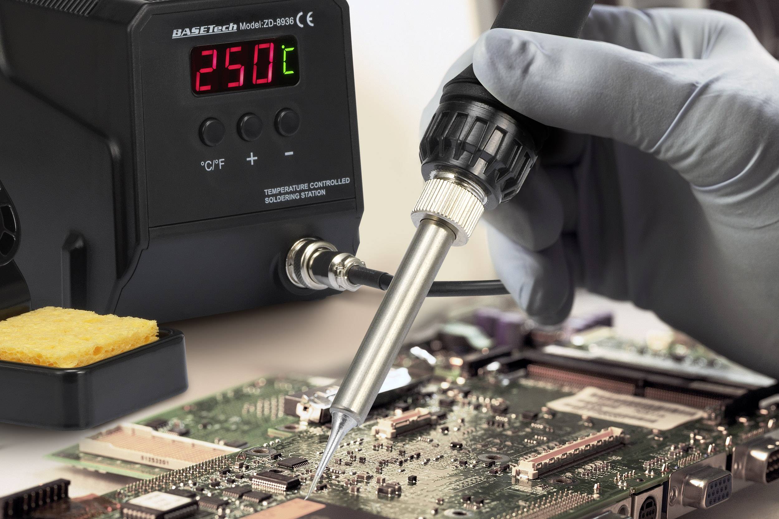Eine Person lötet eine Platine mit einer Lötstation, die auf 250°C eingestellt ist. Ein Schwamm liegt in der Nähe der Station.