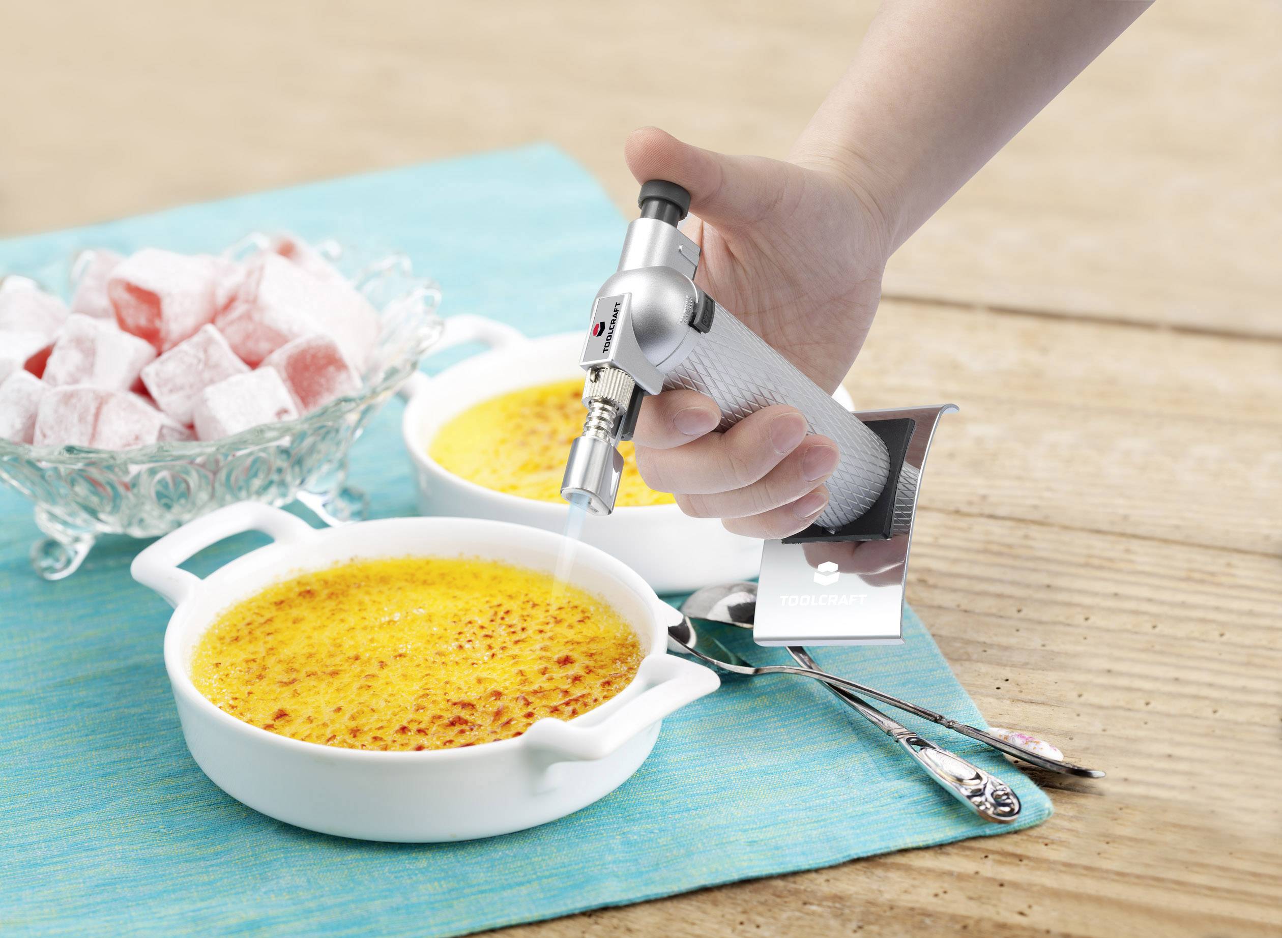 Eine Hand brennt die obere Schicht von zwei Crème Brûlées mit einem Flambierbrenner. Daneben steht eine Schale mit türkischen Delights.