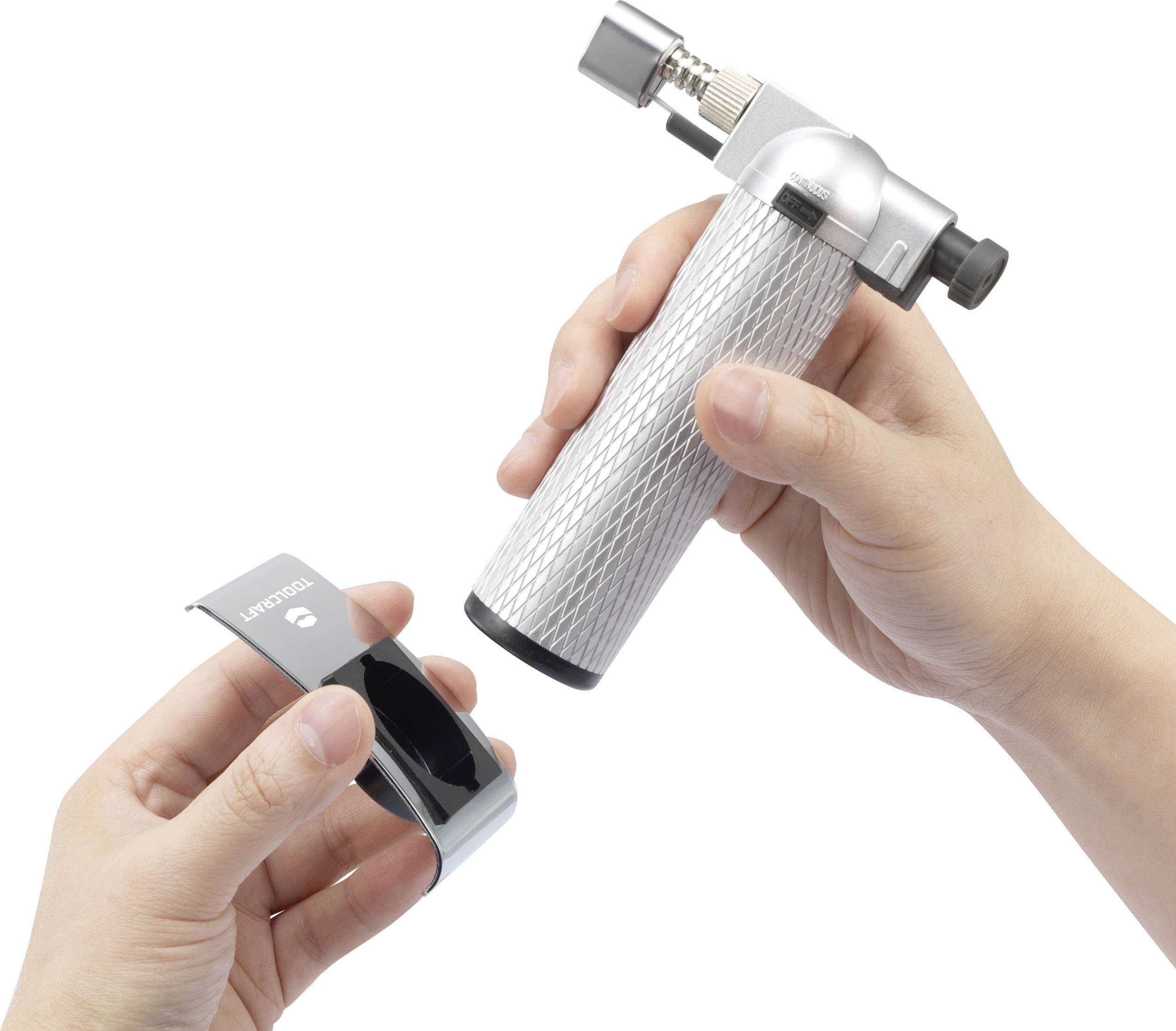 Eine Person hält eine kleine, silberne Lötlampe in der rechten Hand und einen schwarzen, metallischen Ständer in der linken Hand.