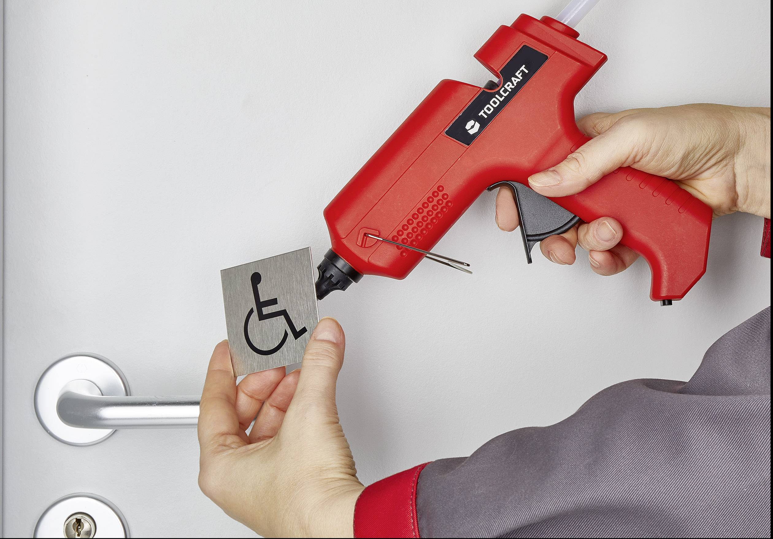 Eine Person hält eine Heißklebepistole und befestigt ein Schild mit einem Rollstuhlsymbol an einer Tür.