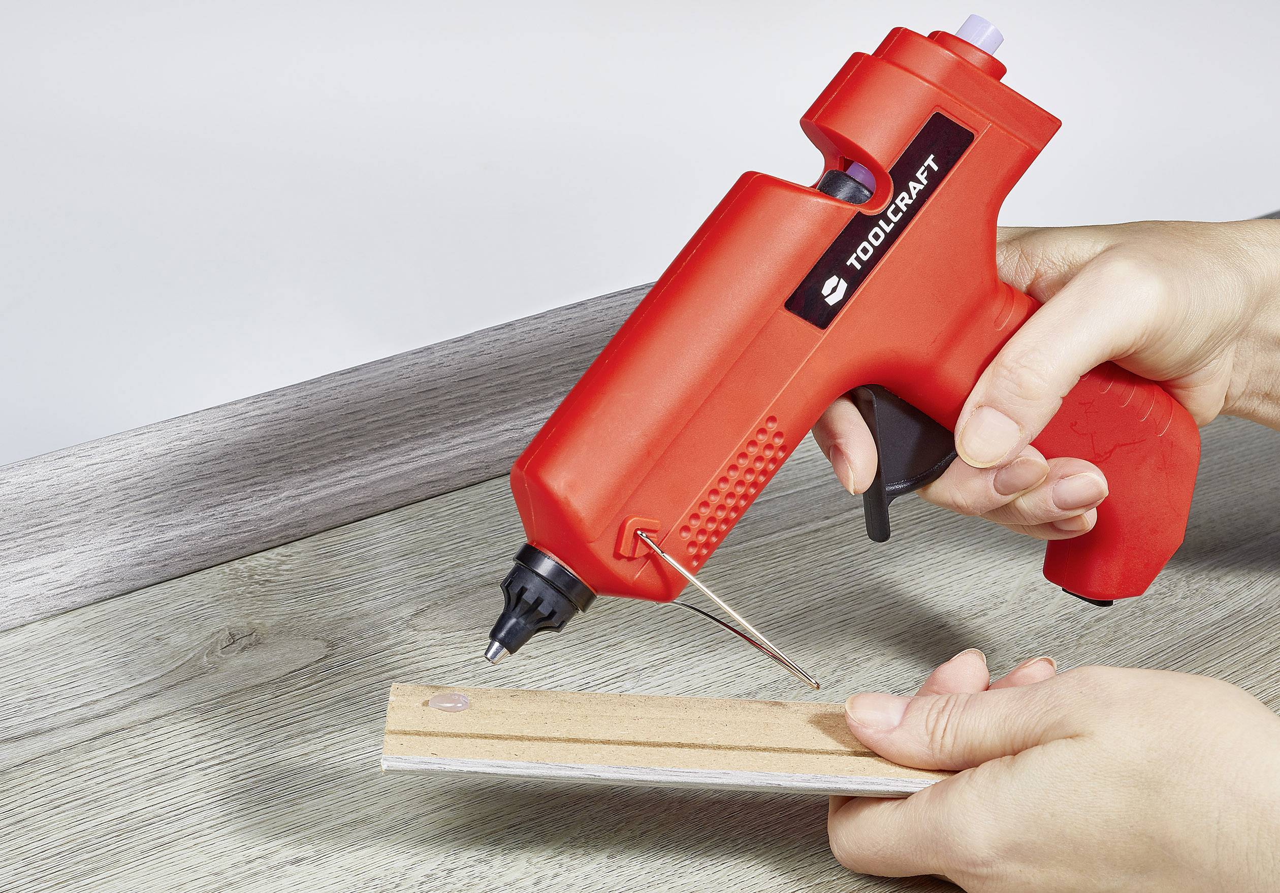 Eine Person verwendet eine rote Heißklebepistole, um ein Holzstück auf einer Tischoberfläche zu kleben.