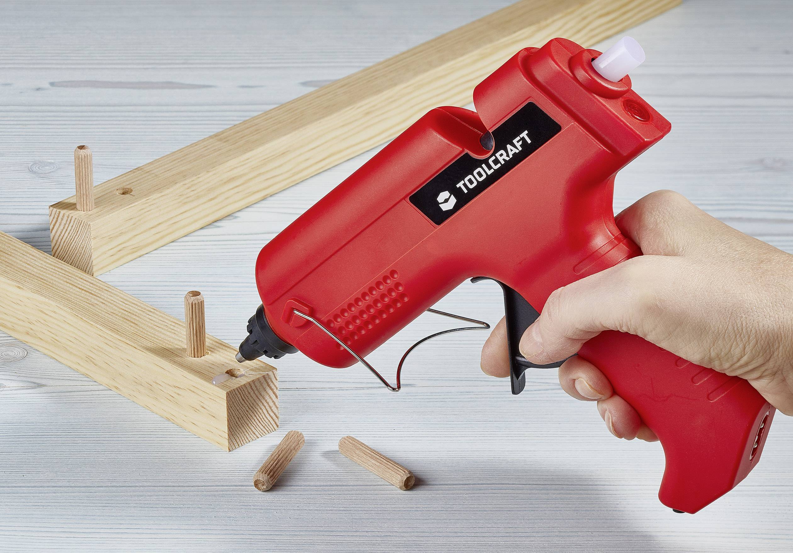 Eine Hand hält eine rote Heißklebepistole, während Holzdübel in Holzteile gesteckt werden, vermutlich für einen Möbelbau.