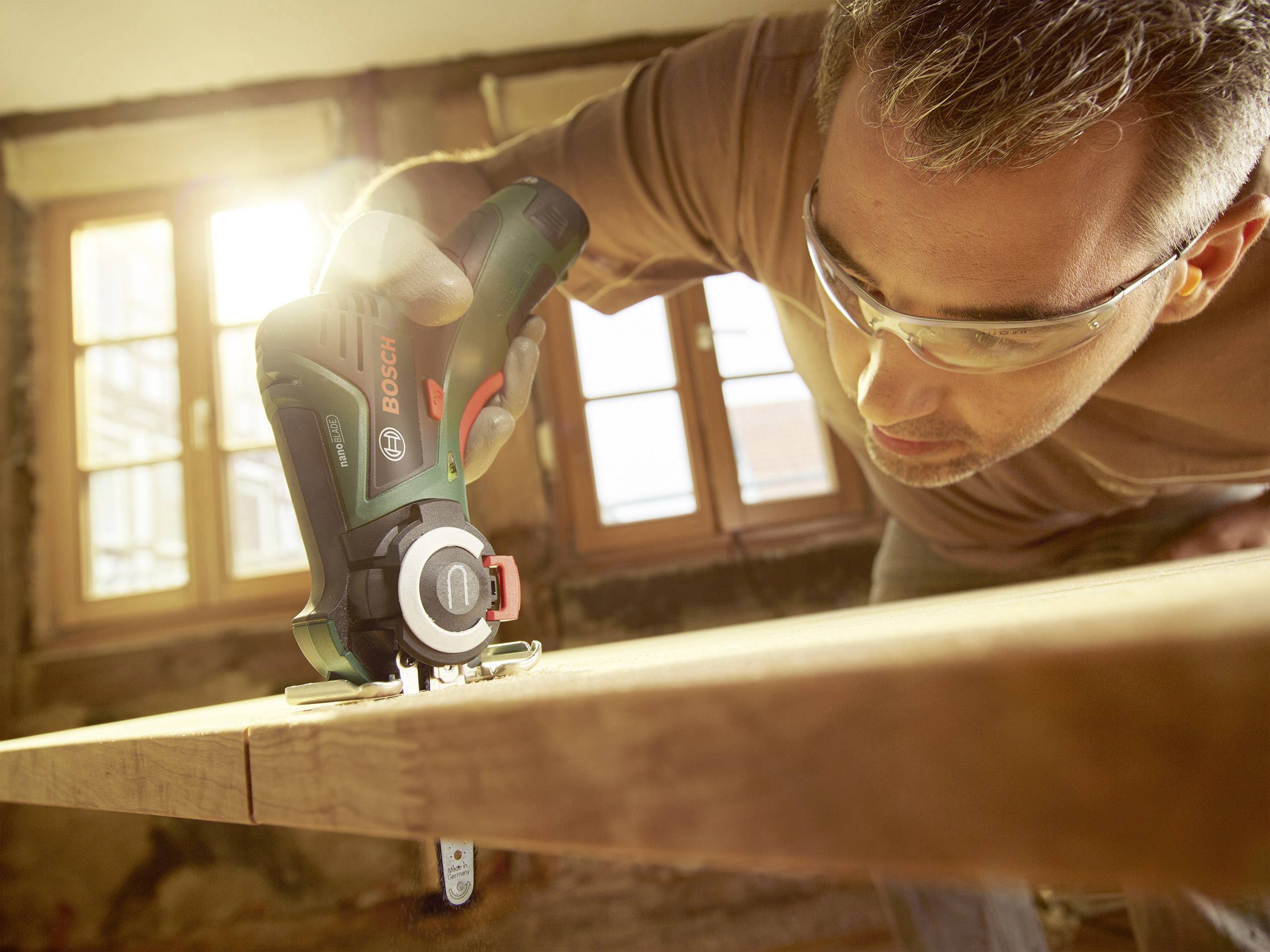 Eine Person benutzt ein elektrisches Handwerkzeug, um Holz zu schneiden. Sonnenlicht fällt durch das Fenster im Hintergrund.