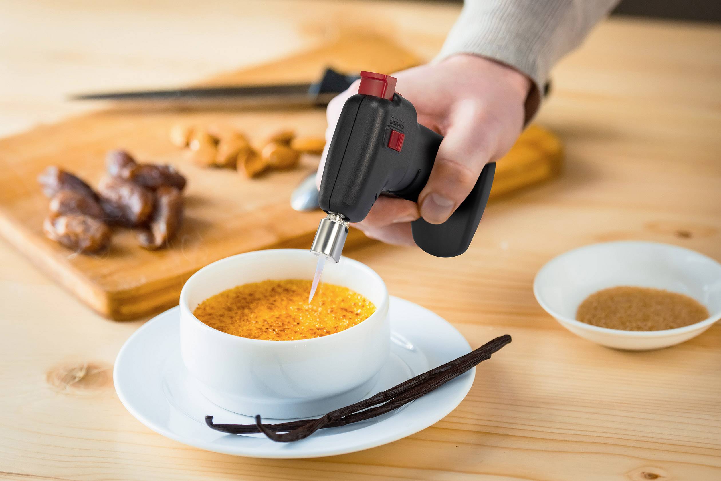 Eine Person karamellisiert mit einem kleinen Gasbrenner die Oberfläche einer Crème brûlée. Im Hintergrund liegen Gewürze und Datteln.