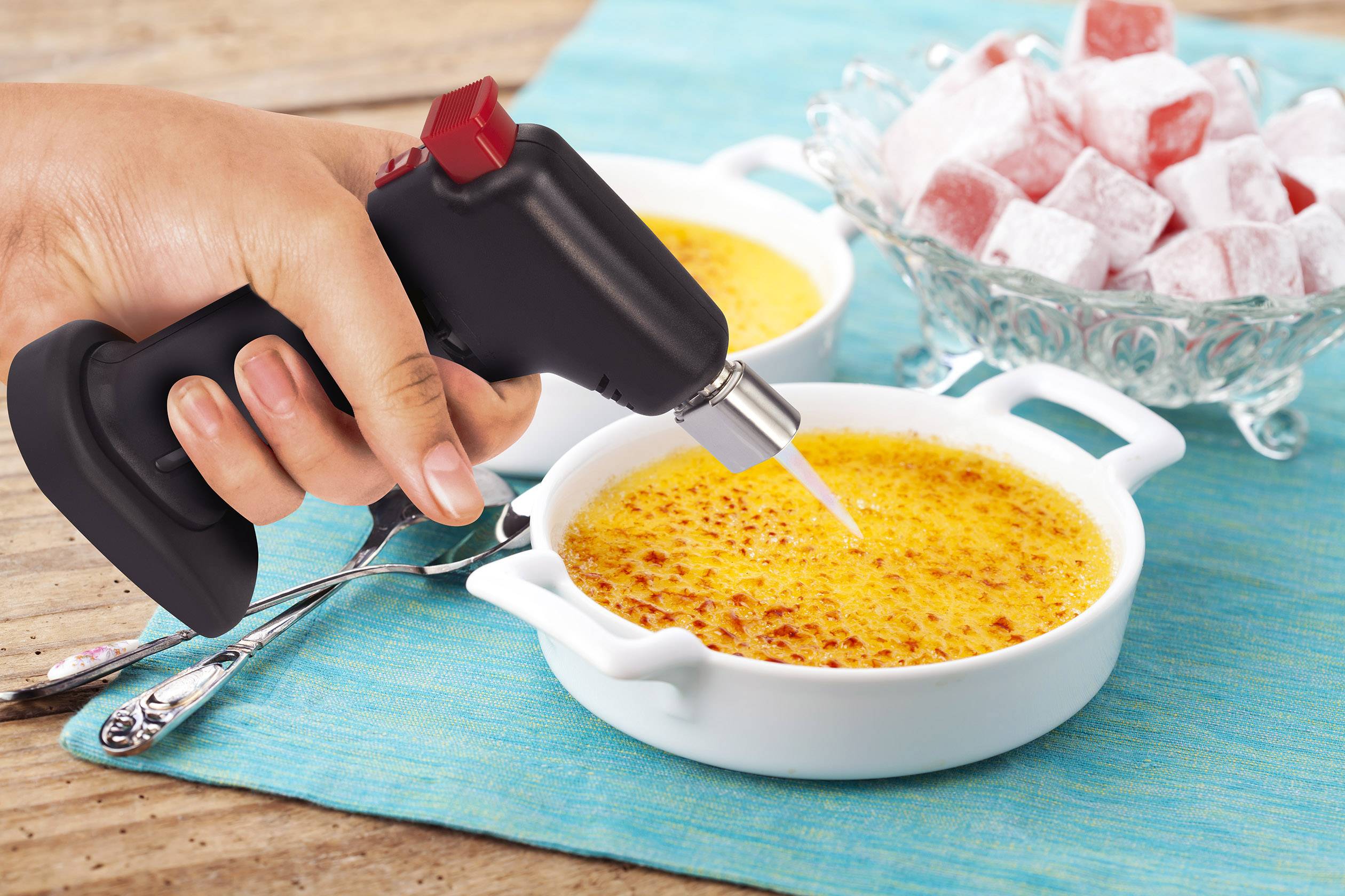 Hand hält Flambierbrenner über Schälchen mit Crème brûlée, daneben eine Schale mit Puderzucker bestäubten Würfeln.