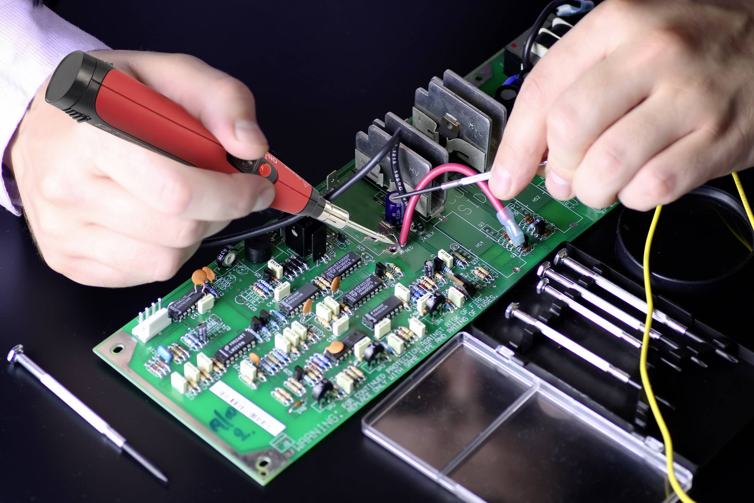 Eine Person lötet mit einem Lötkolben an einer grünen Leiterplatte, umgeben von Werkzeugen.