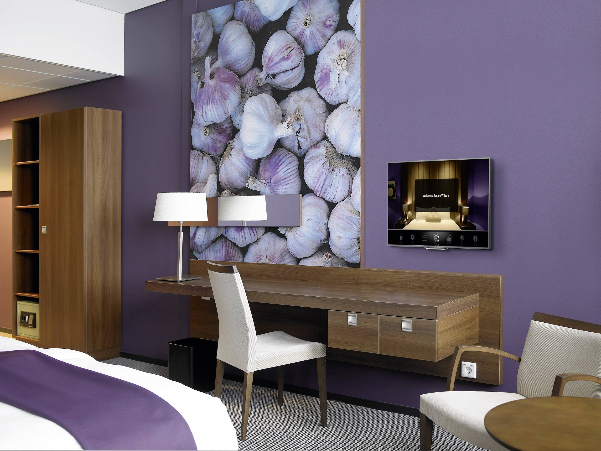 Hotelzimmer mit violetten Wänden, einem Doppelbett, einem Schreibtisch, TV, Lampe und einem großen Knoblauchbild an der Wand.