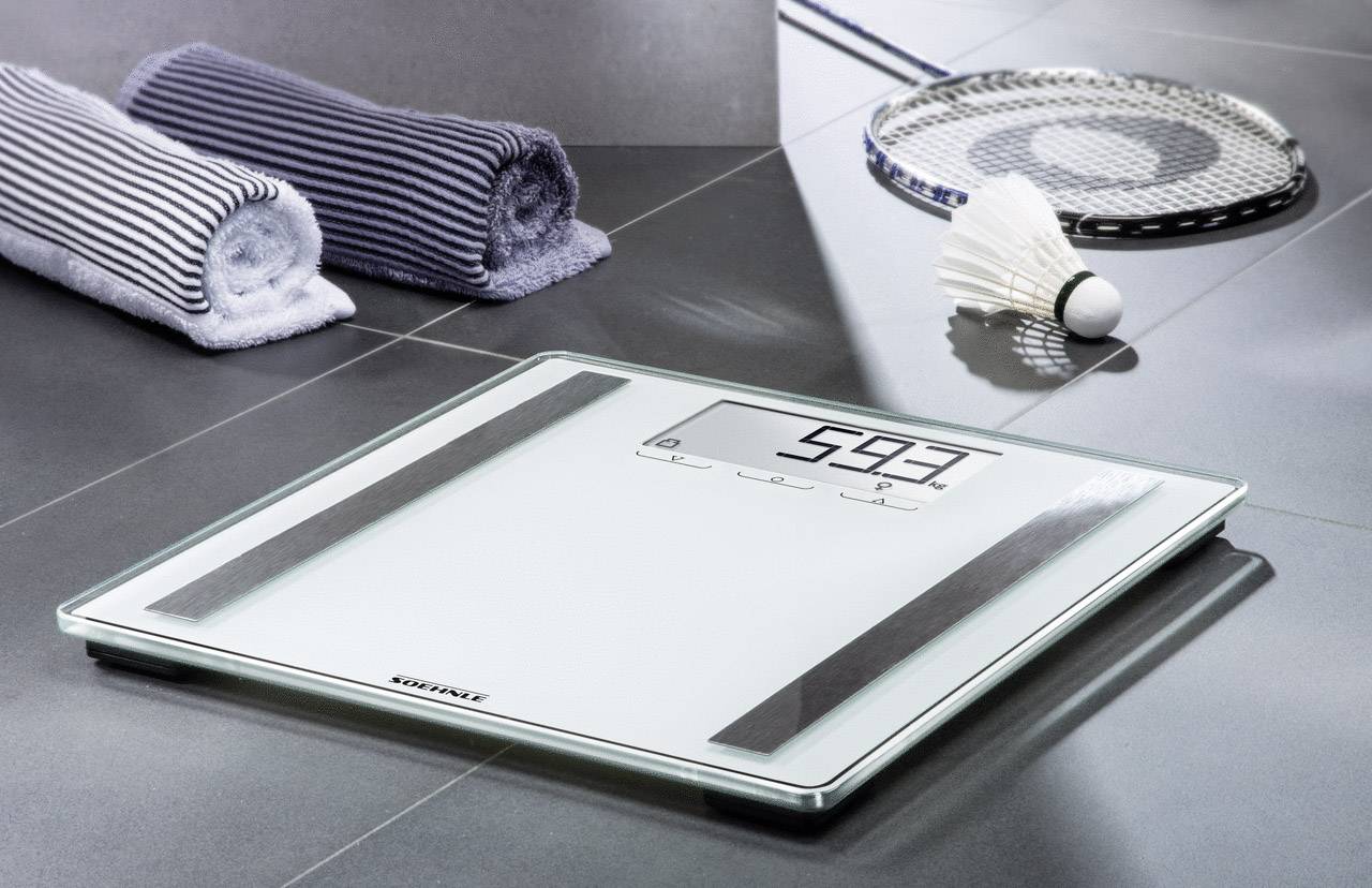 Digitale Waage auf einem Badezimmerboden zeigt 59,3 kg. Daneben liegen zwei gefaltete Handtücher und ein Federball-Schläger mit Federball.