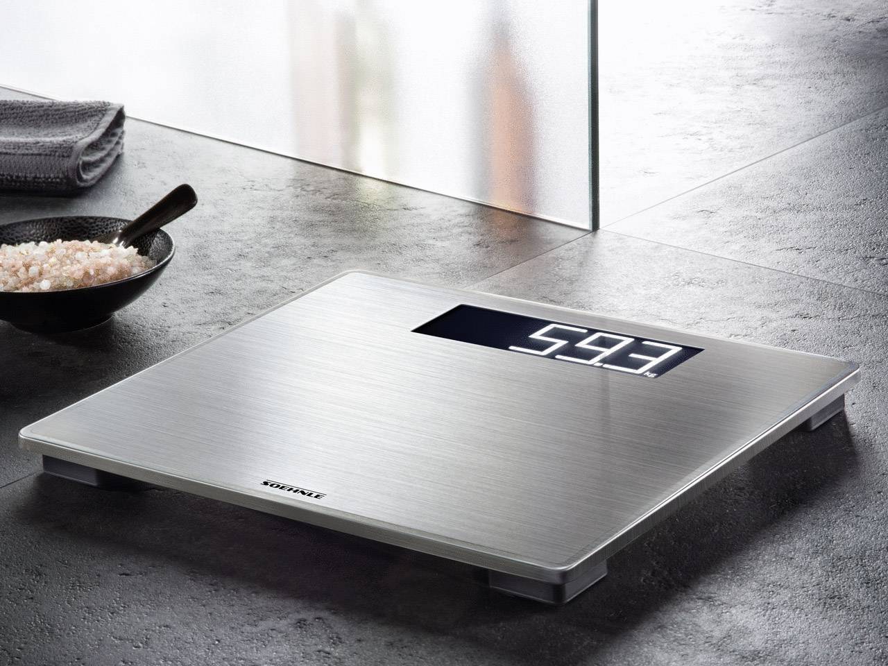 Digitale Waage auf einem grauen Boden zeigt ein Gewicht von 59,3 kg an. Daneben steht eine Schüssel mit rosa Salz und ein Handtuch.