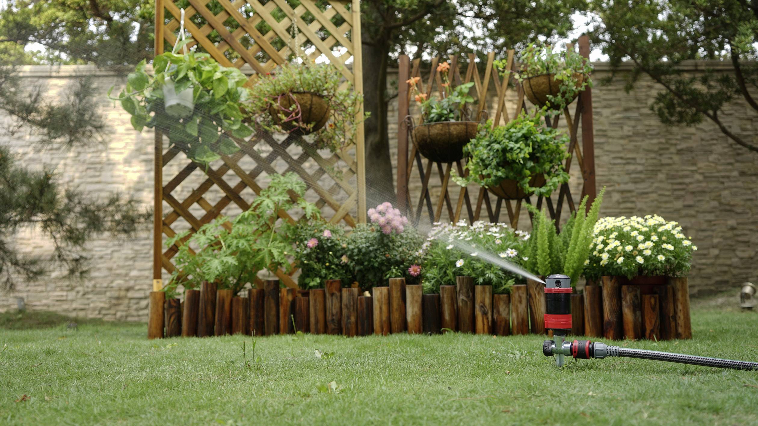 Ein Garten mit Blumenbeet, Holzumrandung und Rankgitter; ein Wasserstrahl bewässert die Pflanzen.