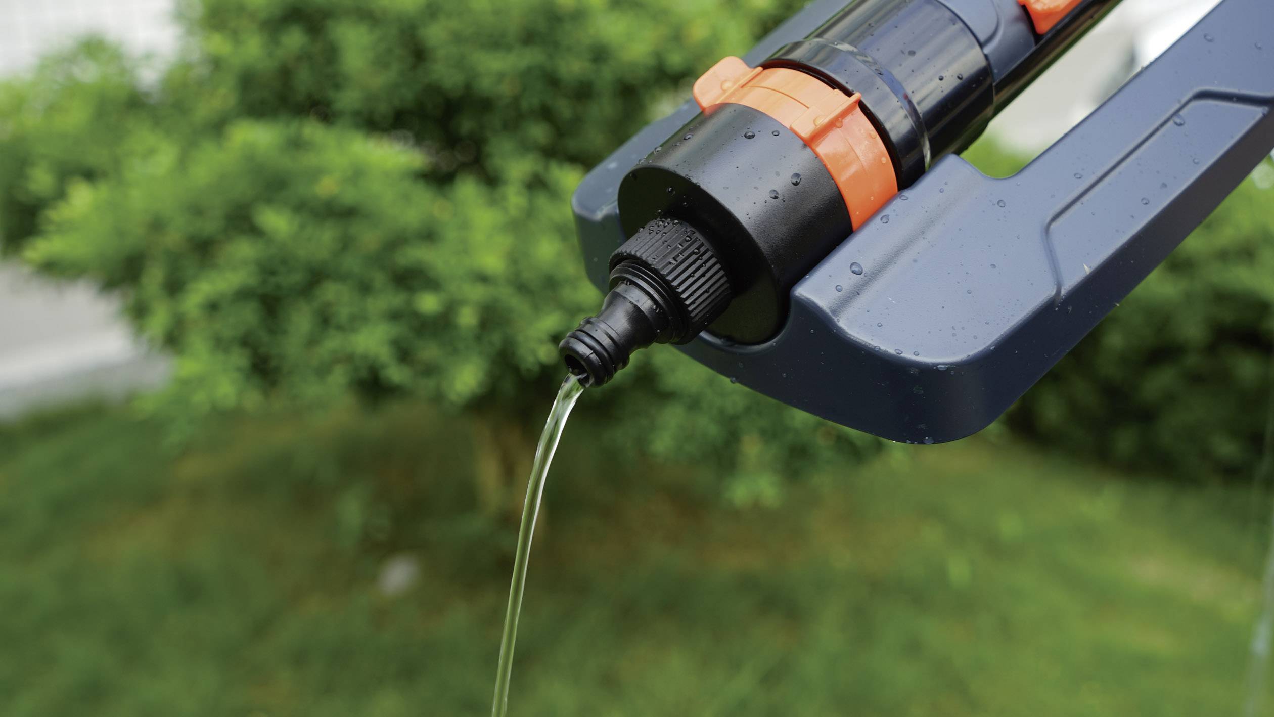 Ein Wasserhahn mit schwarzem und orangefarbenem Design sprüht Wasser auf grüne Pflanzen im Hintergrund, zeigt Bewässerung.