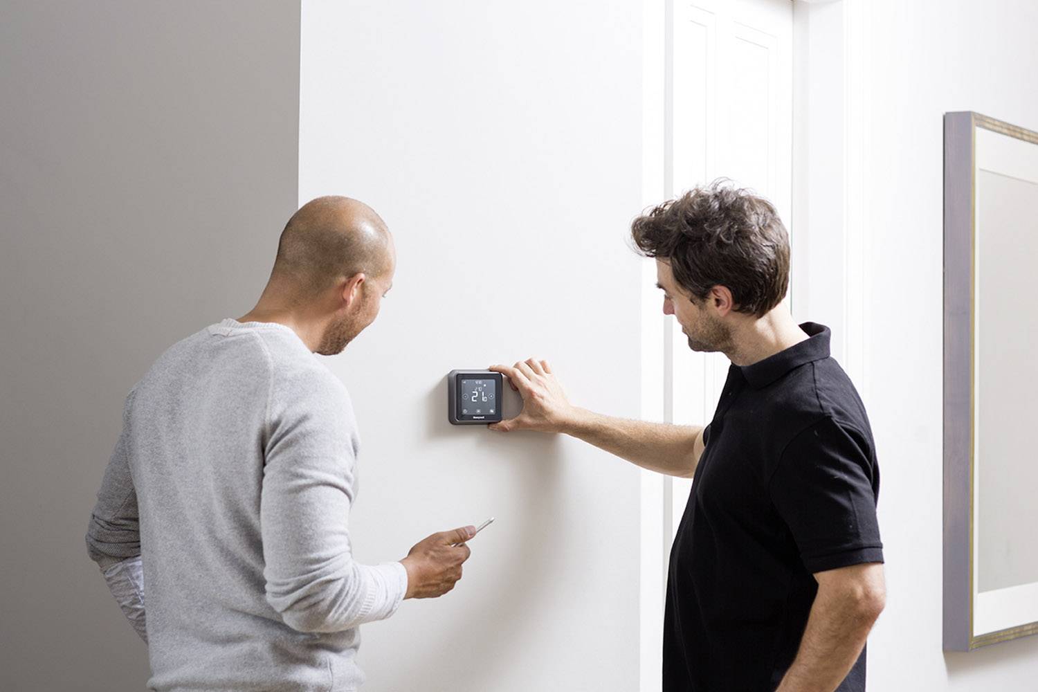 Zwei Männer an einer weißen Wand, einer verstellt ein digitales Thermostat, der andere hält ein Notizbuch.