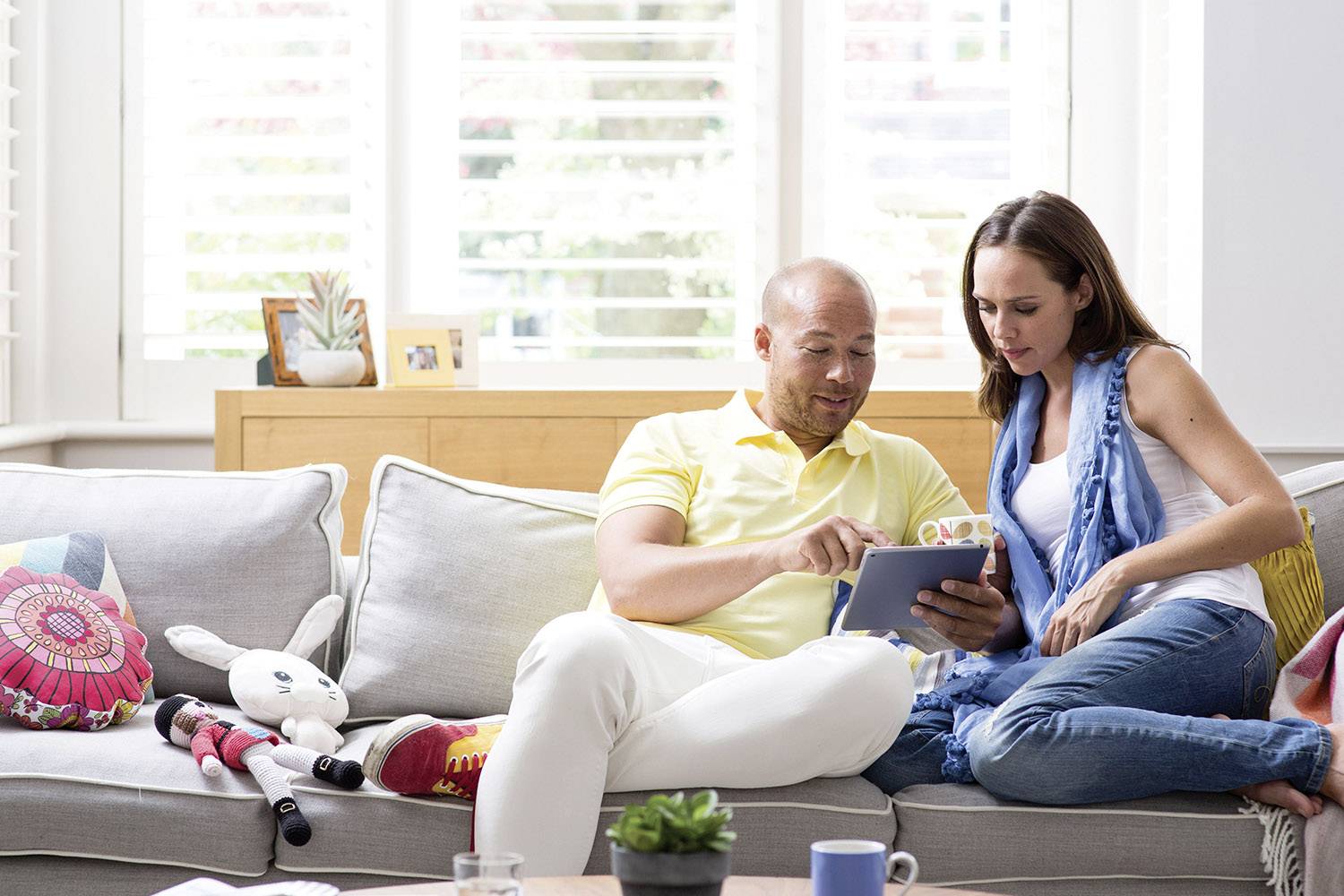 Ein Paar sitzt auf einem Sofa, betrachtet gemeinsam ein Tablet. Im Hintergrund sind ein Fenster und Dekorationen zu sehen.