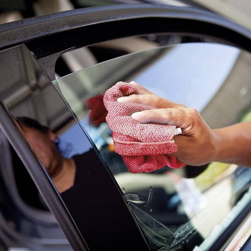 Ein rotes Tuch wischt eine Autoscheibe. Eine Person hält das Tuch, sichtbar durch einen Teil ihres Arms, im Sonnenlicht.