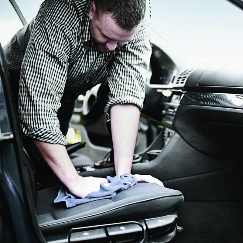 Eine Person reinigt den Fahrersitz eines Autos mit einem blauen Tuch. Der Innenraum ist modern und aufgeräumt, mit Fokus auf Sauberkeit.