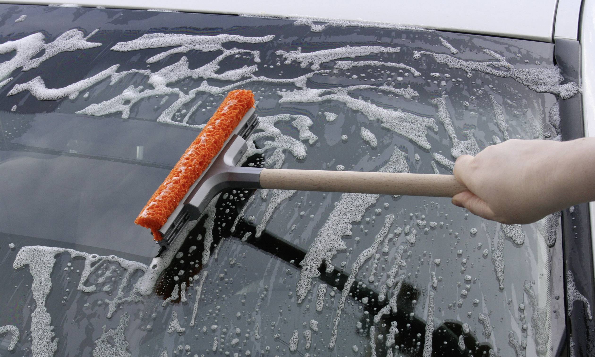 Eine Hand reinigt die Windschutzscheibe eines Autos mit einem Abzieher und Seifenwasser. Der Hintergrund zeigt Teile des Fahrzeugs.