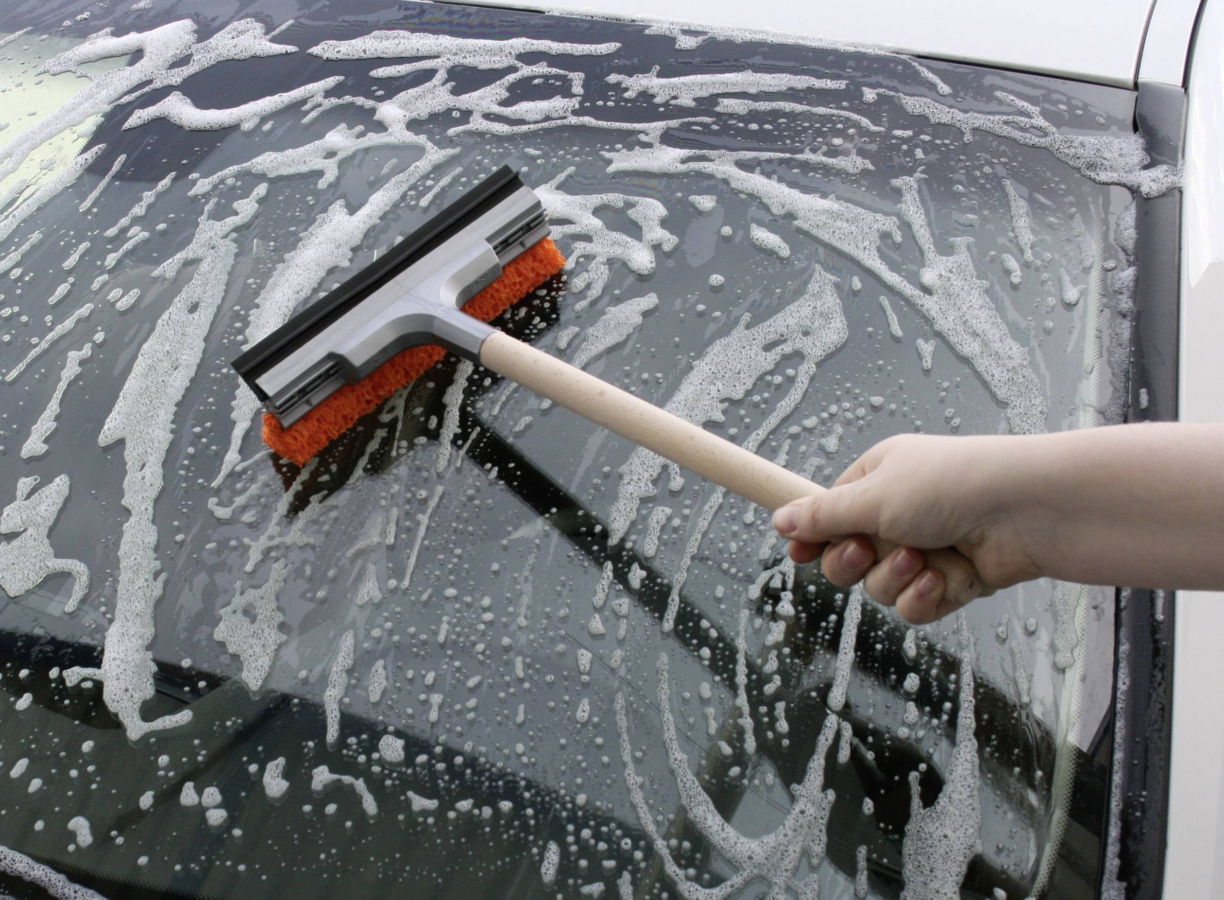 Eine Hand reinigt mit einem Scheibenwischer und Seifenwasser die Windschutzscheibe eines Autos.