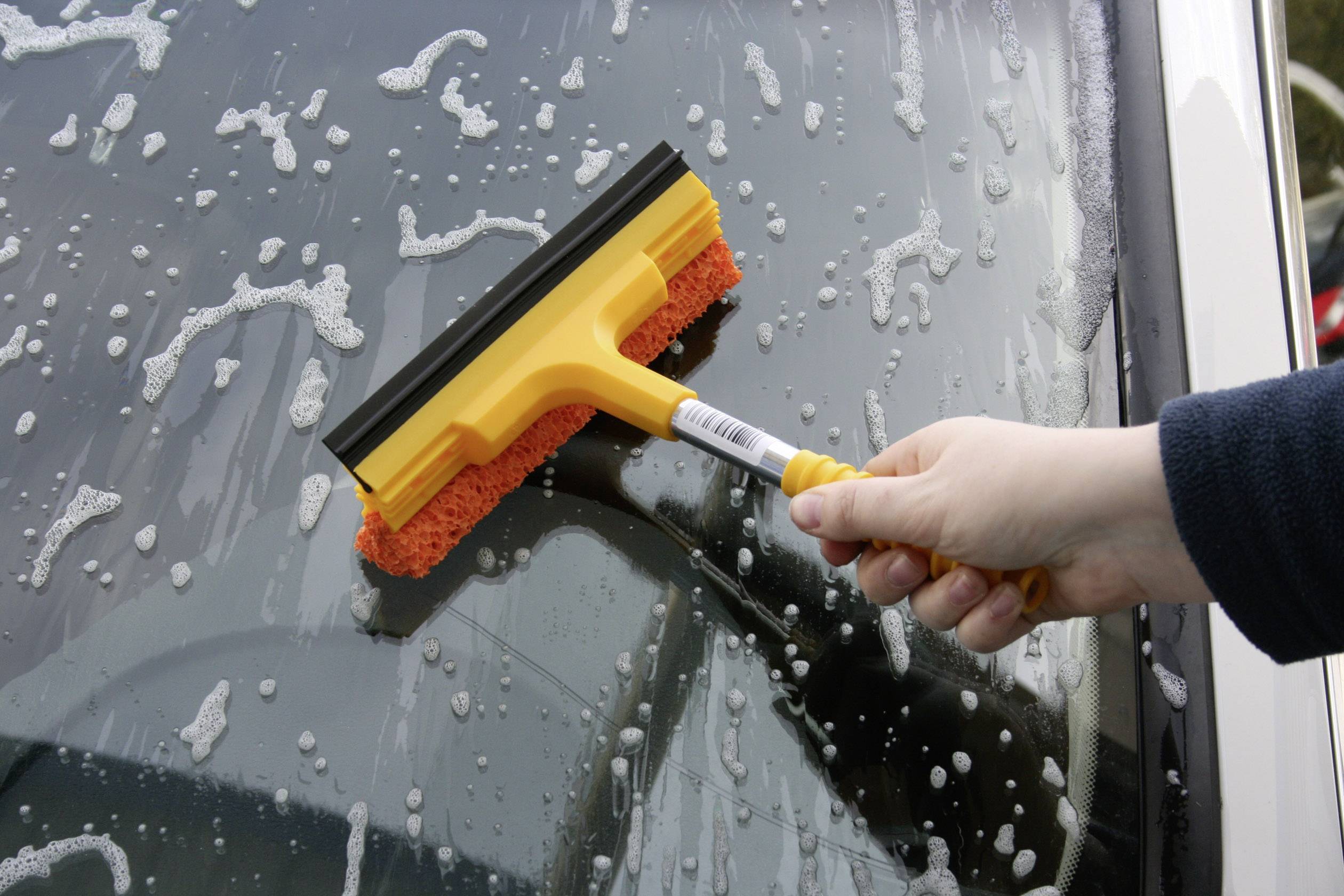 Eine Person reinigt die Außenseite eines Autofensters mit einem orangefarbenen Abzieher, entfernt Seifenwasser und Schaum.