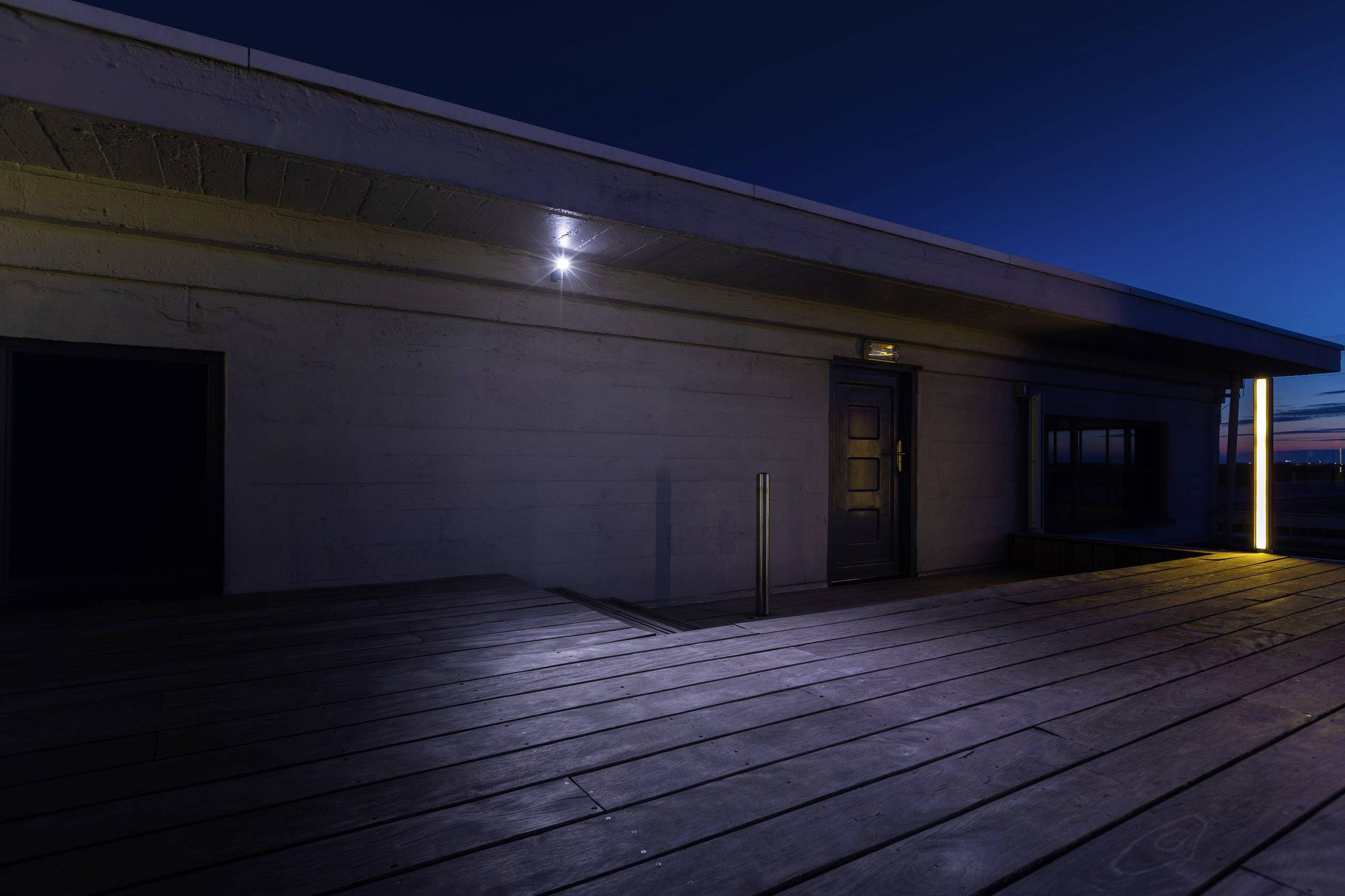Ein modernes Gebäude bei Nacht mit Holzdeck und schwacher Beleuchtung. Im Hintergrund ist ein schwacher Streifen des Sonnenuntergangs zu sehen.