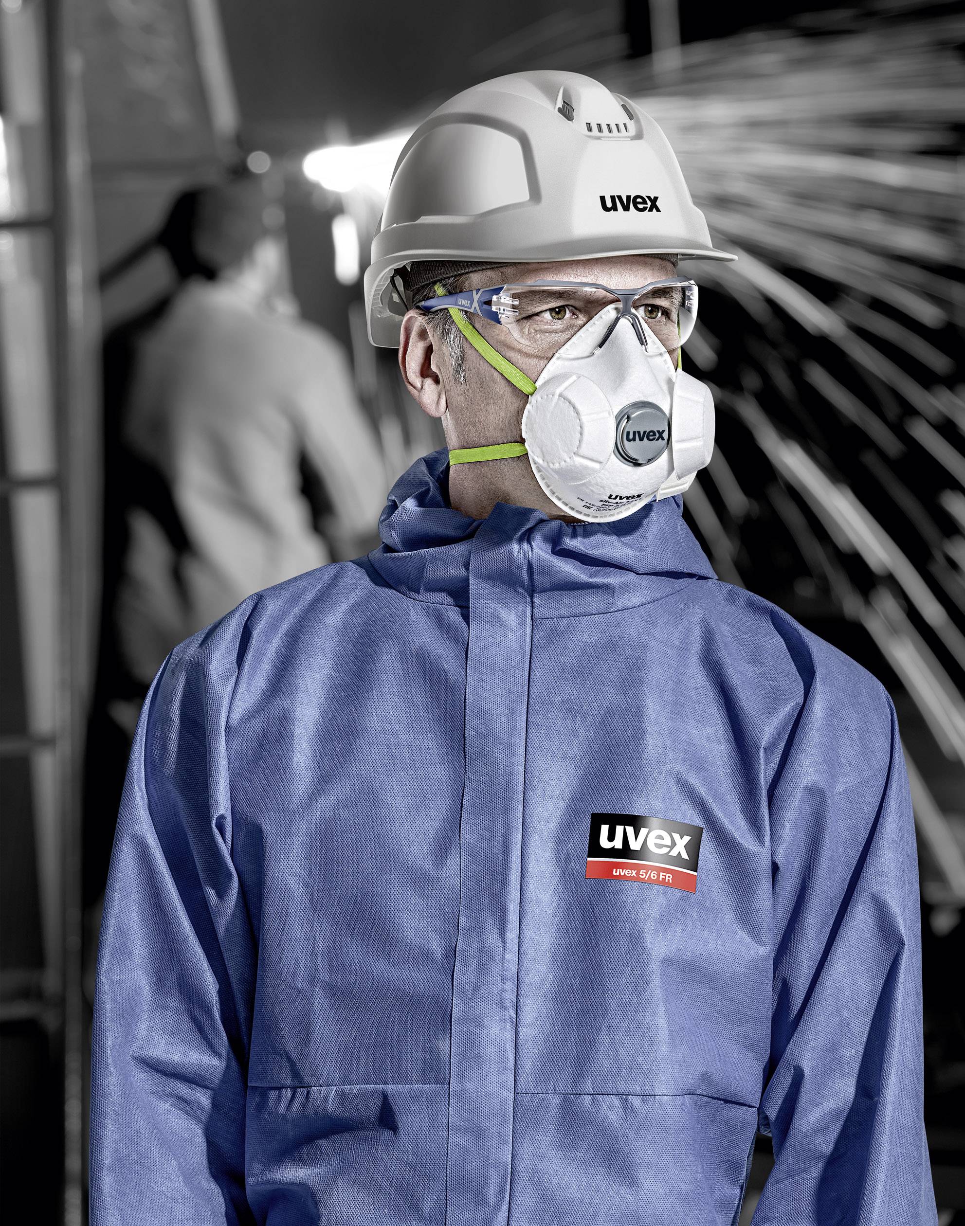 Person in blauer Arbeitskleidung, mit Helm und Schutzmaske, steht in einer industriellen Umgebung mit Funken im Hintergrund.