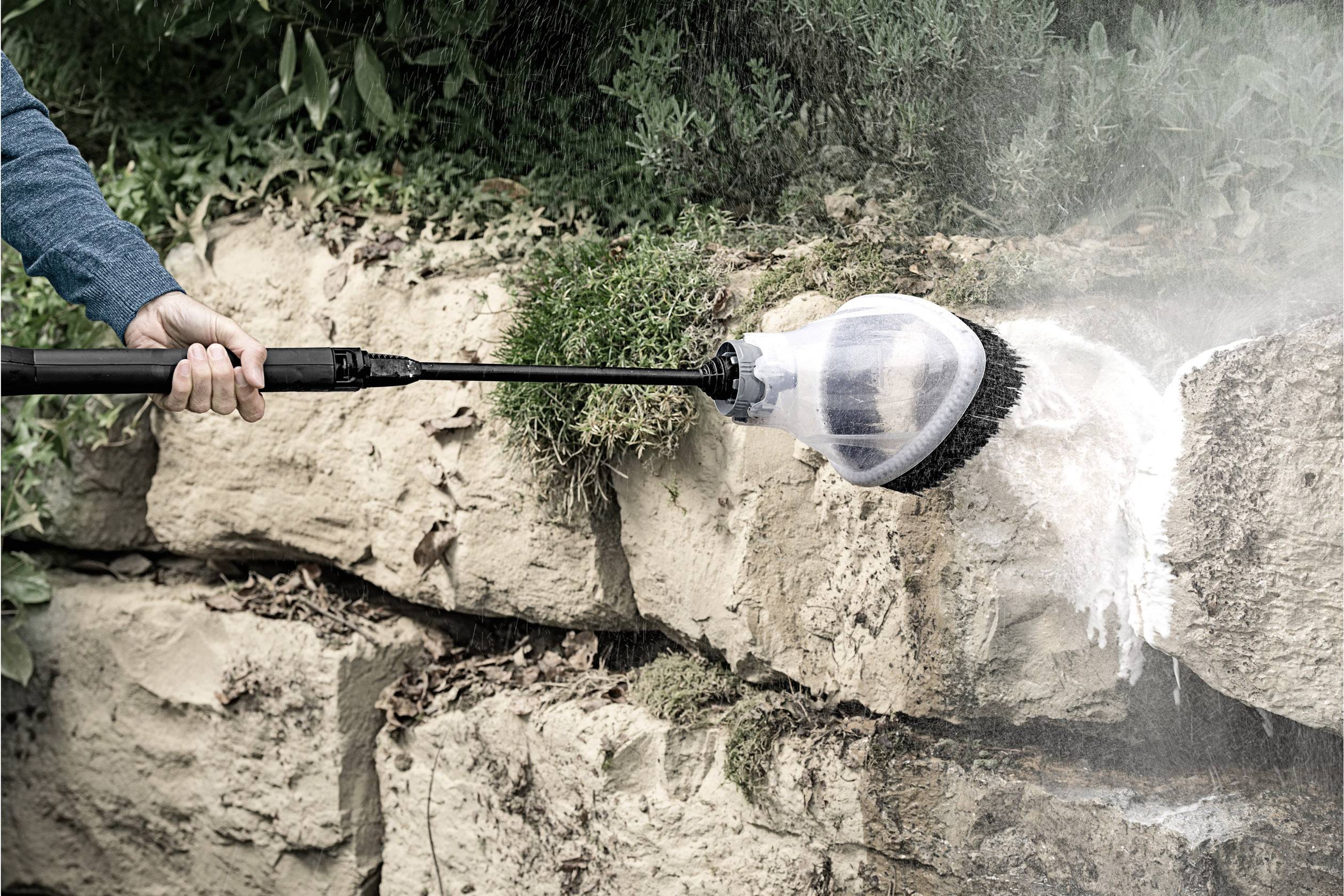 Ein Druckreiniger wäscht Felsen. Eine Rundbürste entfernt Schmutz von einer steinernen Oberfläche. Im Hintergrund Pflanzen.