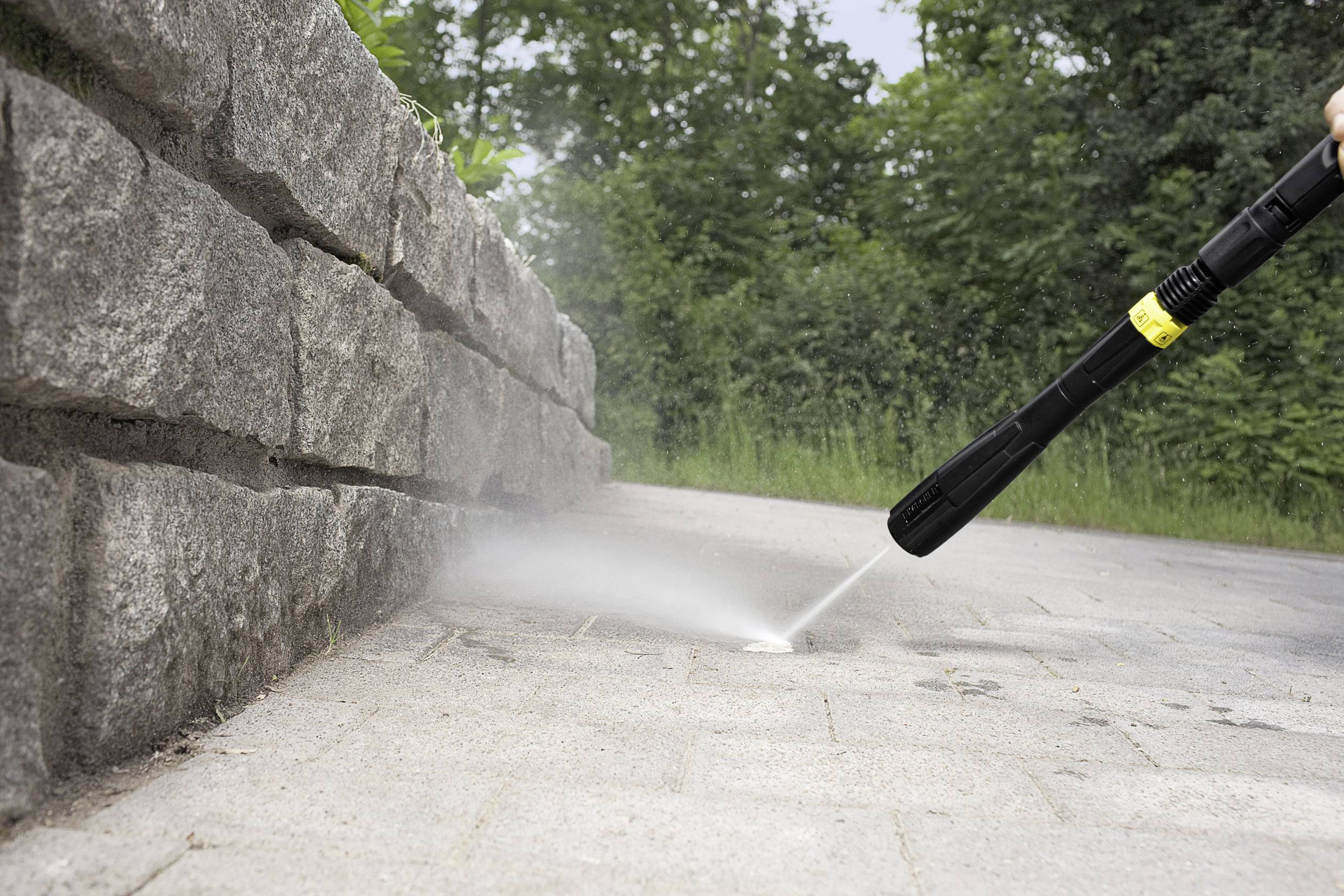 Eine Person reinigt mit einem Hochdruckreiniger die Fugen eines gepflasterten Weges neben einer Steinmauer im Freien.