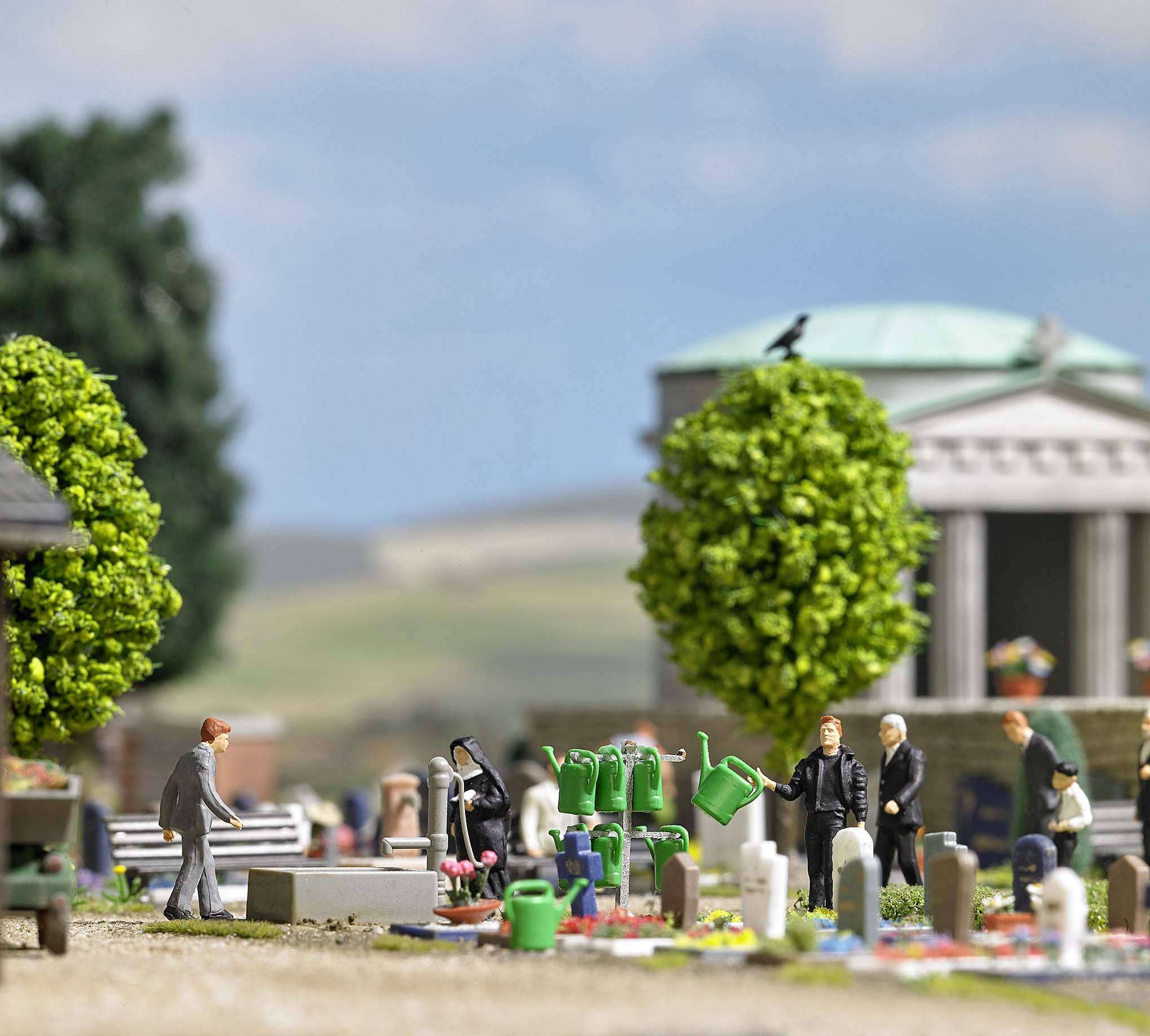 Personen auf einem Miniaturfriedhof mit Grabsteinen, Bäumen und Gieskannen. Im Hintergrund sind ein Tempelgebäude und Landschaft sichtbar.