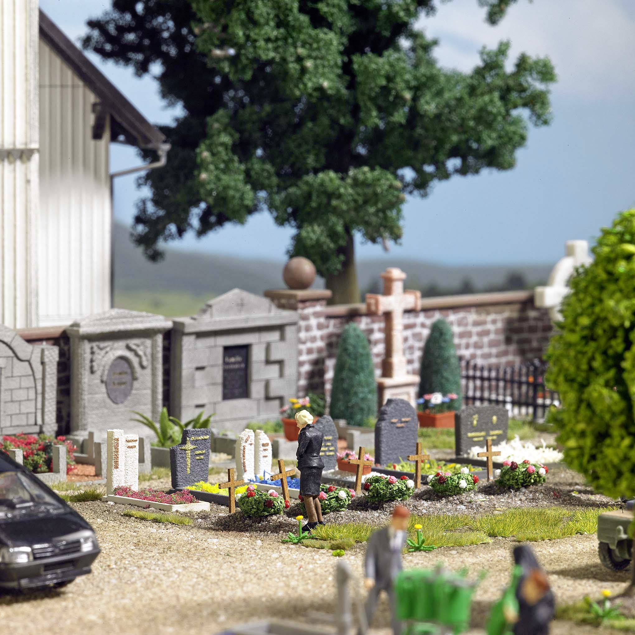 Ein Miniaturfriedhof mit Grabsteinen, einem Betenden, Bäumen und einem Auto im Vordergrund, umgeben von grüner Landschaft und blauem Himmel.