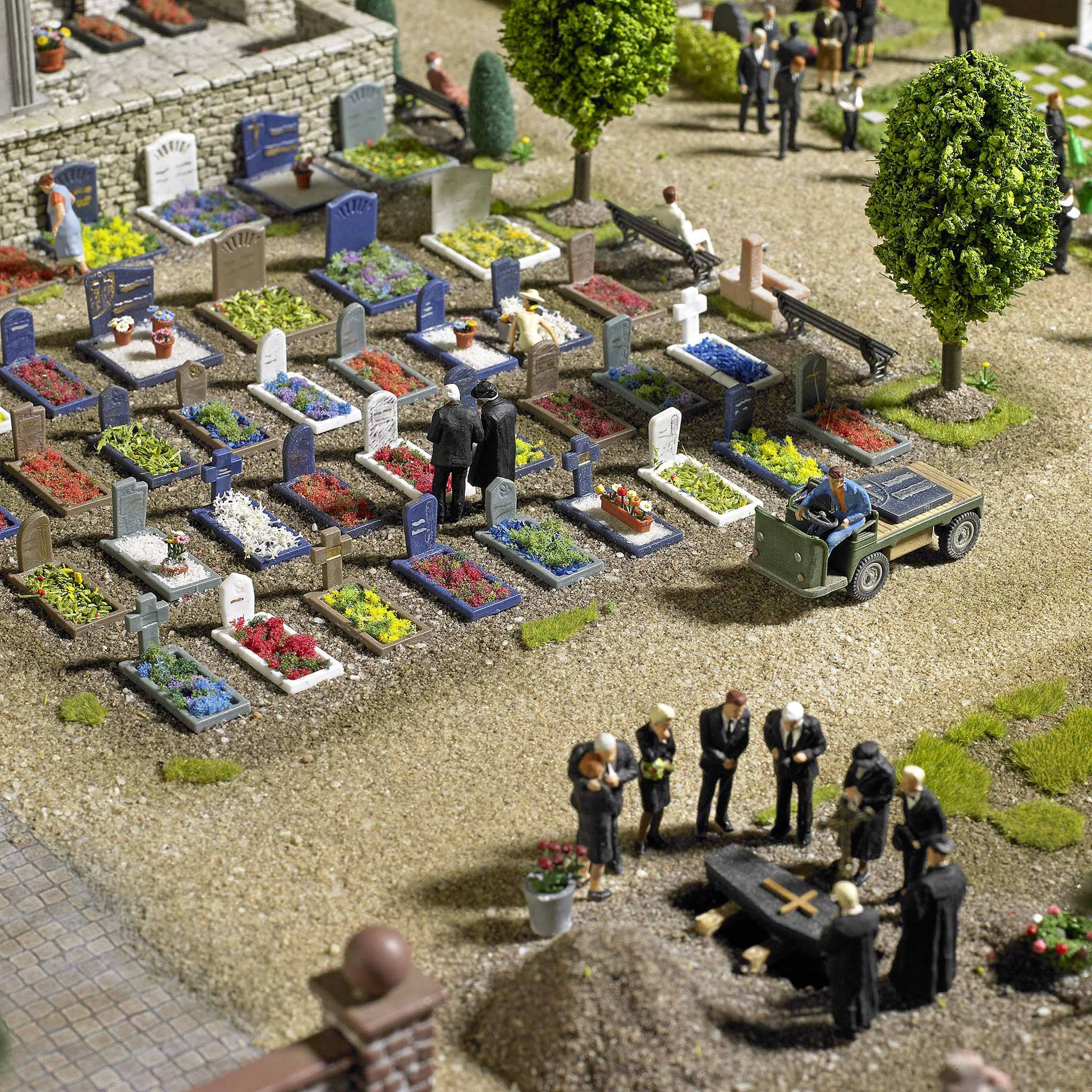 Miniatur-Friedhofsszene mit Figuren bei einer Bestattung, mehrere Gräber, ein grüner Wagen. Bäume und Blumen säumen den Weg.