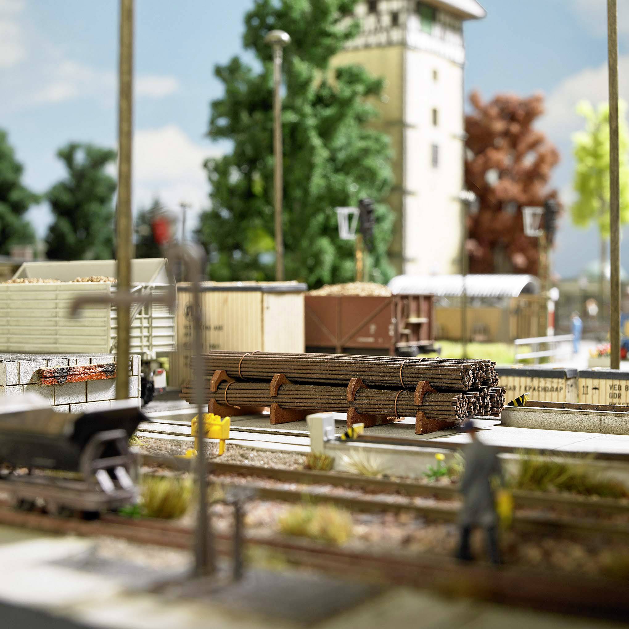 Modelleisenbahnlandschaft mit Schienen, Waggons und Gebäuden, umgeben von Bäumen und Zäunen. Im Hintergrund ein mehrstöckiges Gebäude.