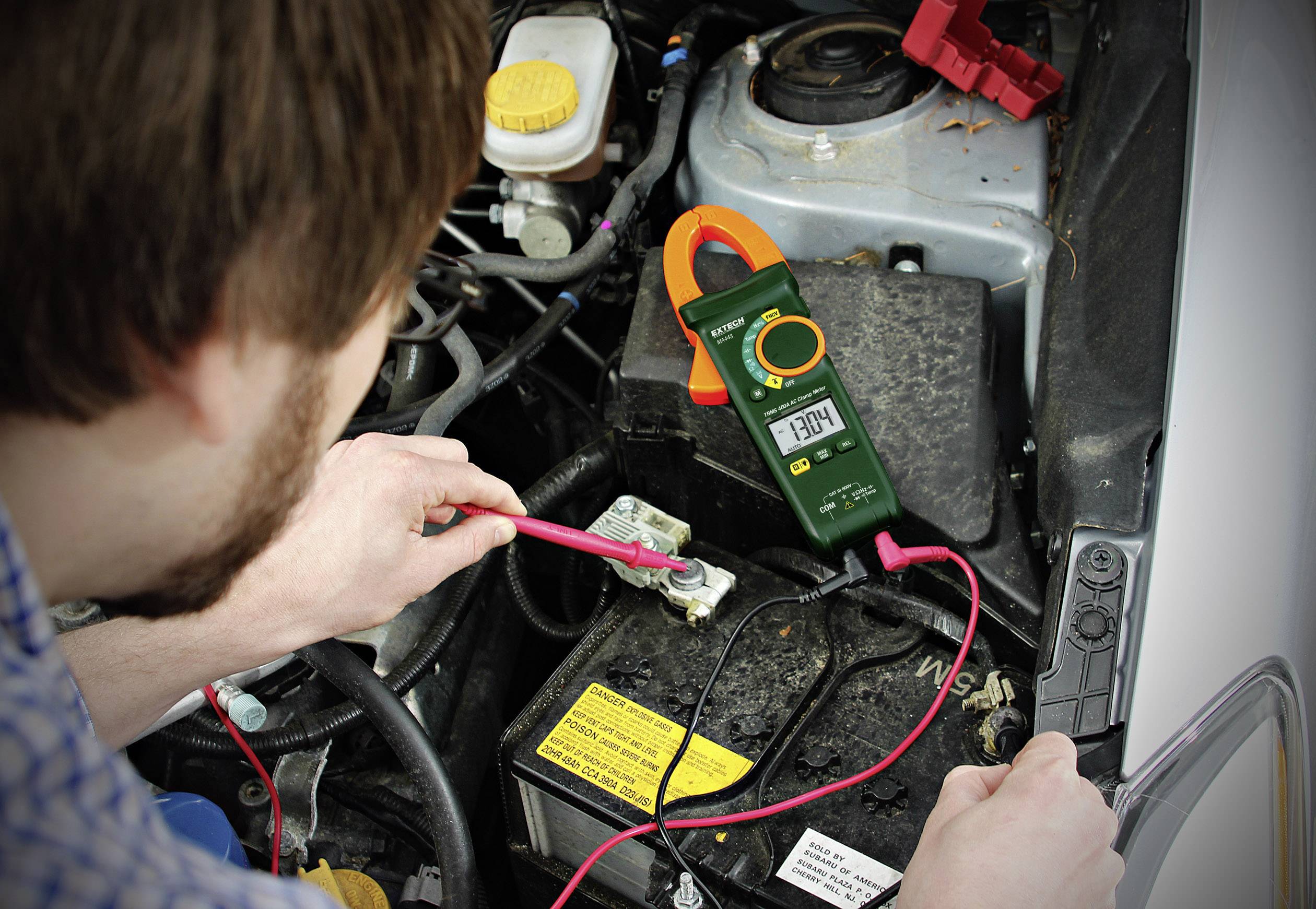 Ein Mann misst die Spannung einer Autobatterie mit einem Multimeter. Das Display zeigt 13,08 Volt an, während rote Kabel an den Polen angeschlossen sind.