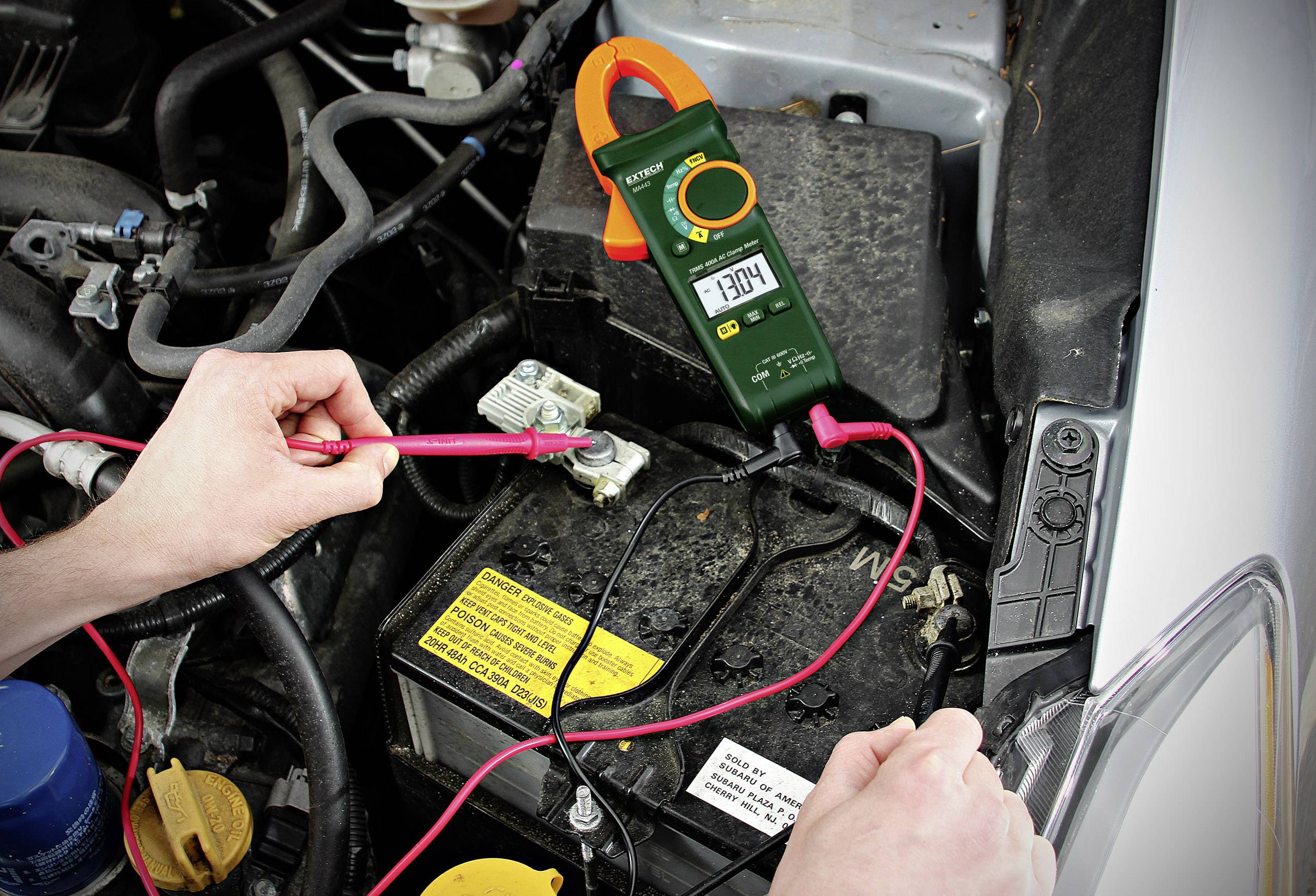 Eine Person misst die Spannung einer Autobatterie mit einem Multimeter, das 13,04 Volt anzeigt. Kabel und Motorraum sind sichtbar.