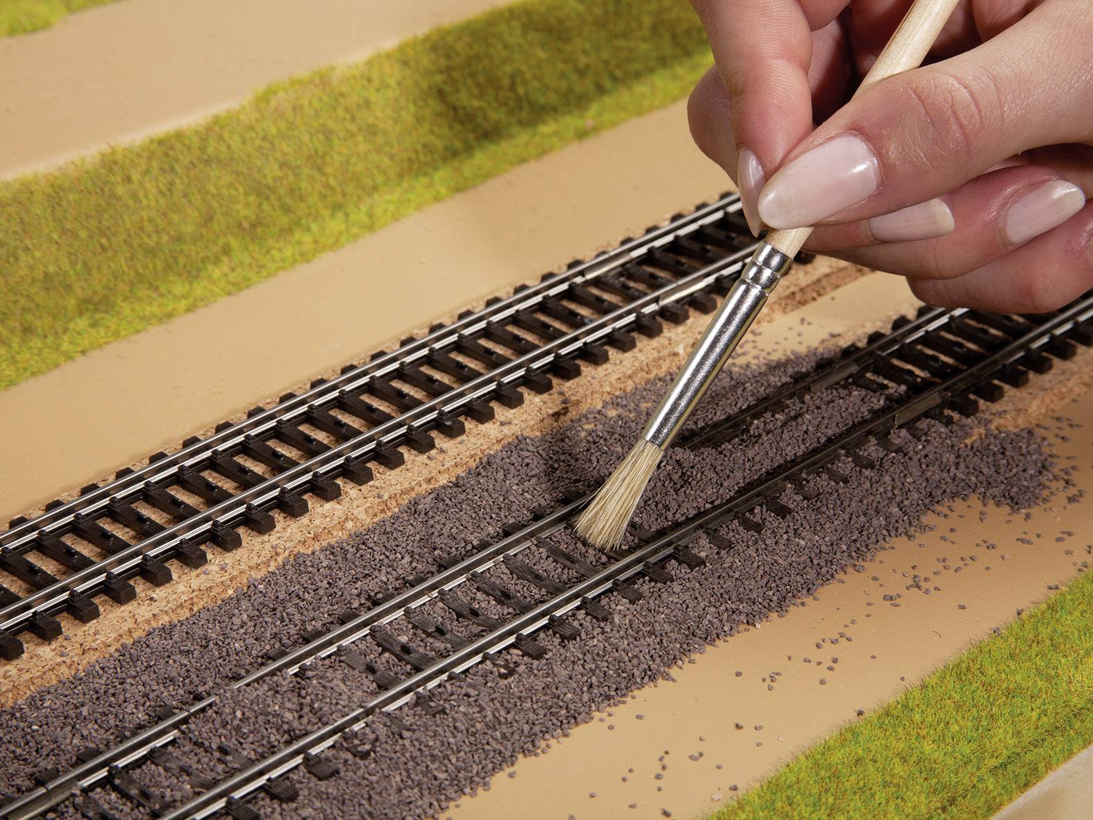 Eine Hand pinselt feine Steine auf ein Modellbahngleis, um eine realistische Landschaft zu gestalten.