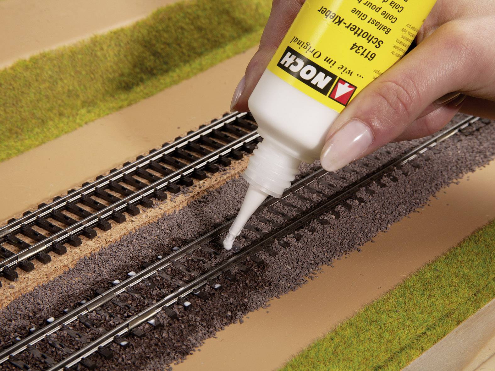 Hand trägt mit einer Flasche Klebstoff auf Modellbahngleise auf. Die Szene zeigt eine Modellbauarbeit auf einer Landschaftsplatte.