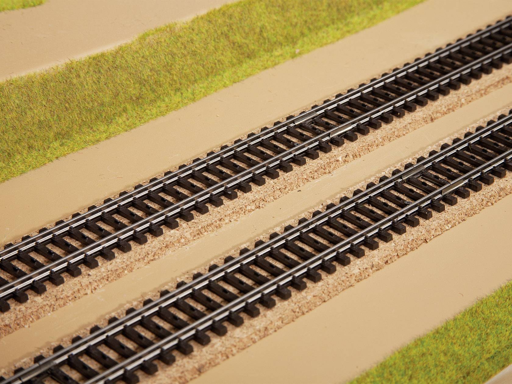 Zwei parallele Eisenbahnschienen auf sandigem Untergrund mit grasbewachsenen Seitenstreifen, in einem Modellbahn-Layout dargestellt.
