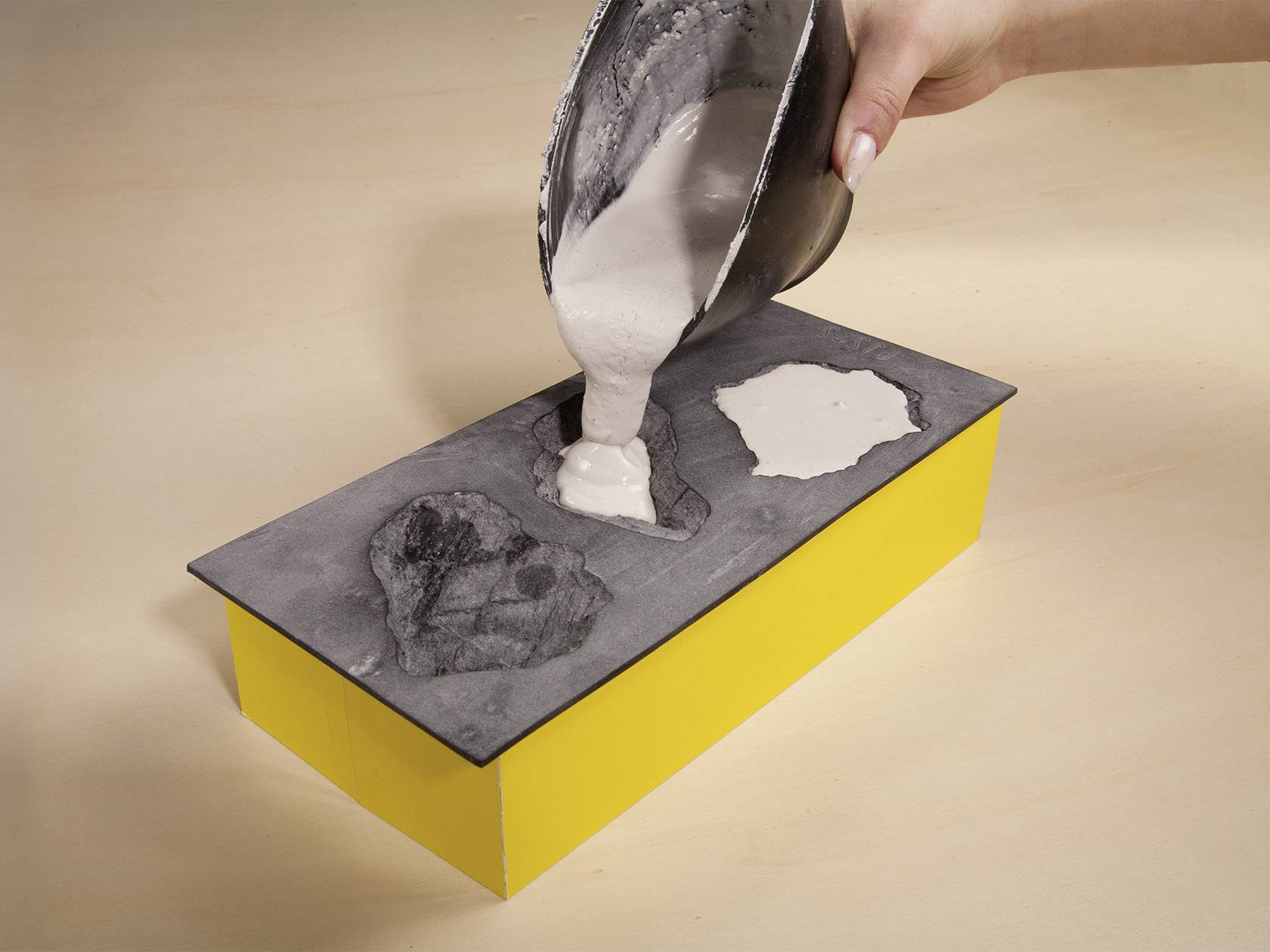 Eine Hand gießt eine flüssige Substanz aus einer Metallschale in eine rechteckige Form mit gelben Seiten auf einem Tisch.
