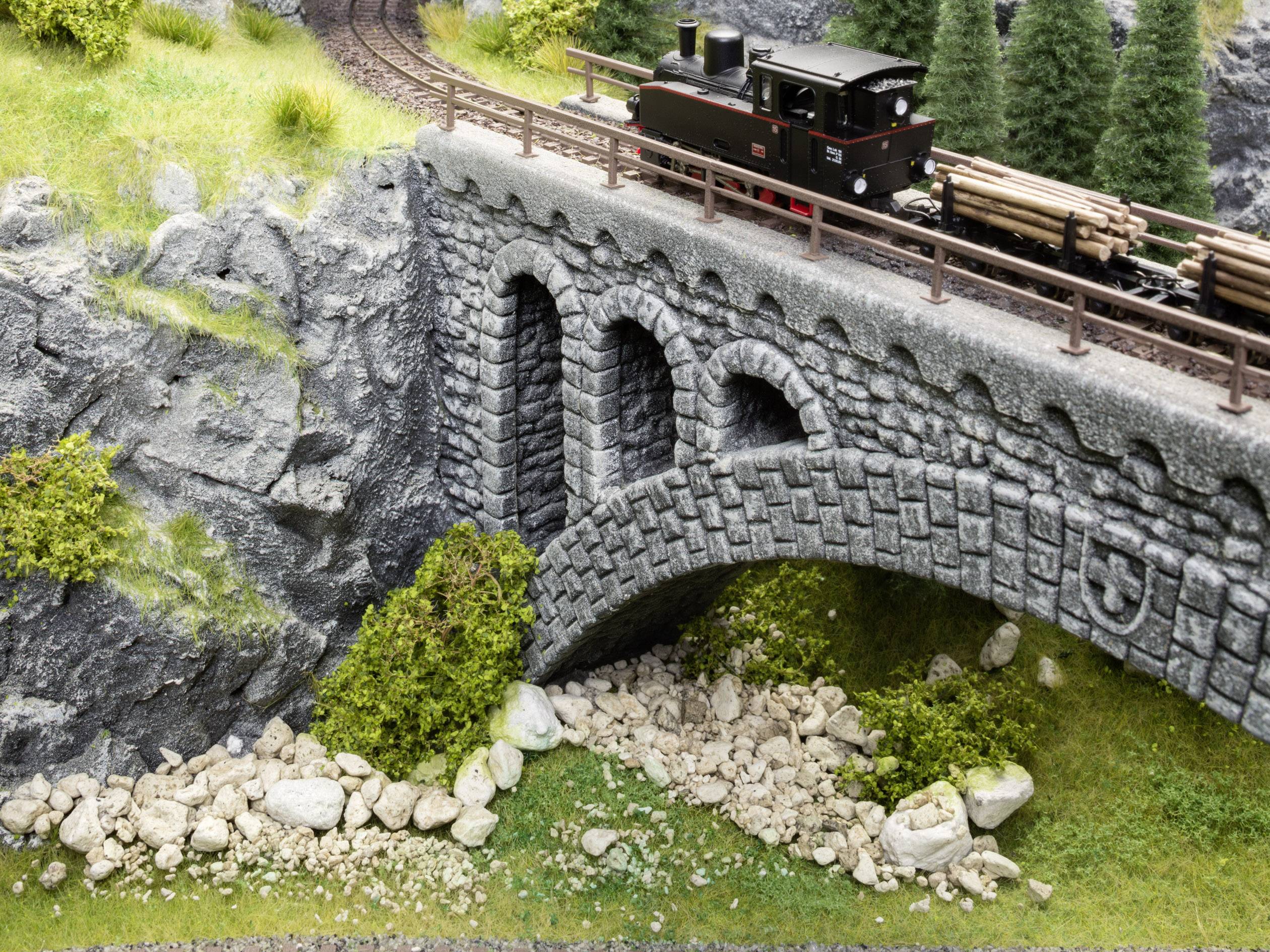 Eine Miniatureisenbahn fährt über eine steinerne Brücke in einer bergigen Landschaft mit Felsen und Bäumen.