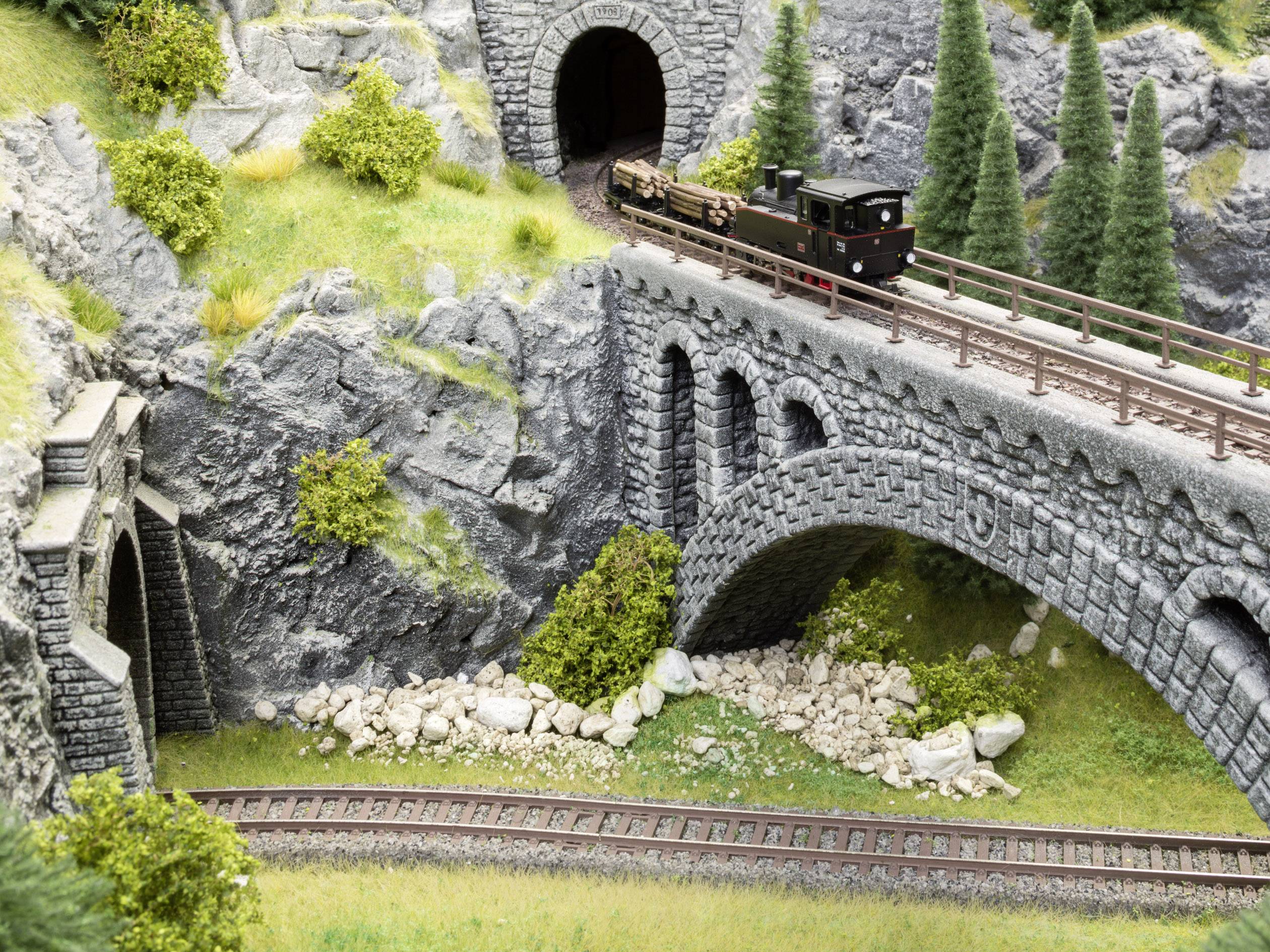 Modelleisenbahnlandschaft mit Steinbrücke, Tunnel und kleinem Zug. Der Zug fährt über die Brücke, umgeben von Felsen und Bäumen.