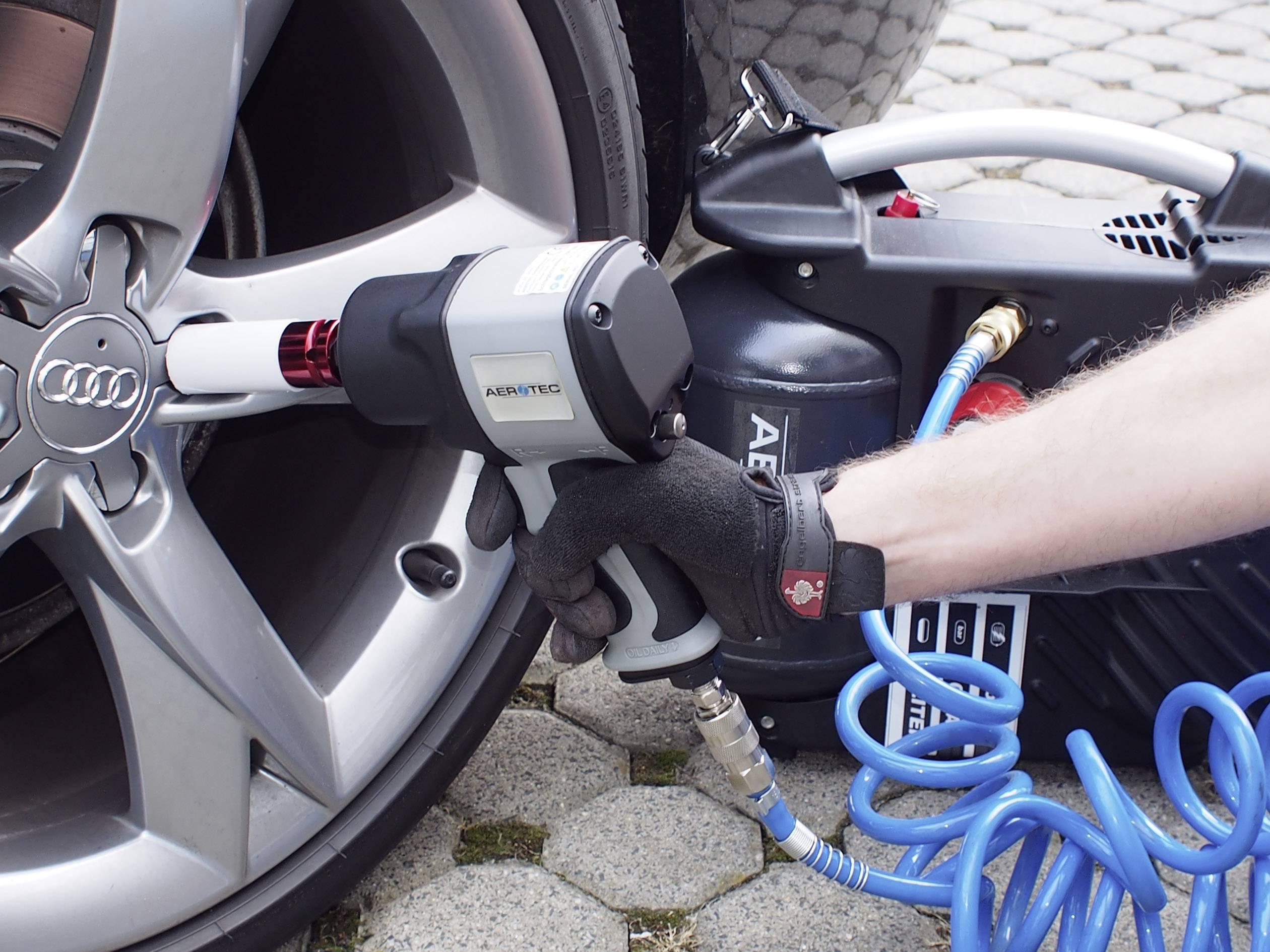 Eine Person wechselt ein Rad eines Autos mit einem Druckluftschrauber; ein Reifen und Werkzeug sind sichtbar.